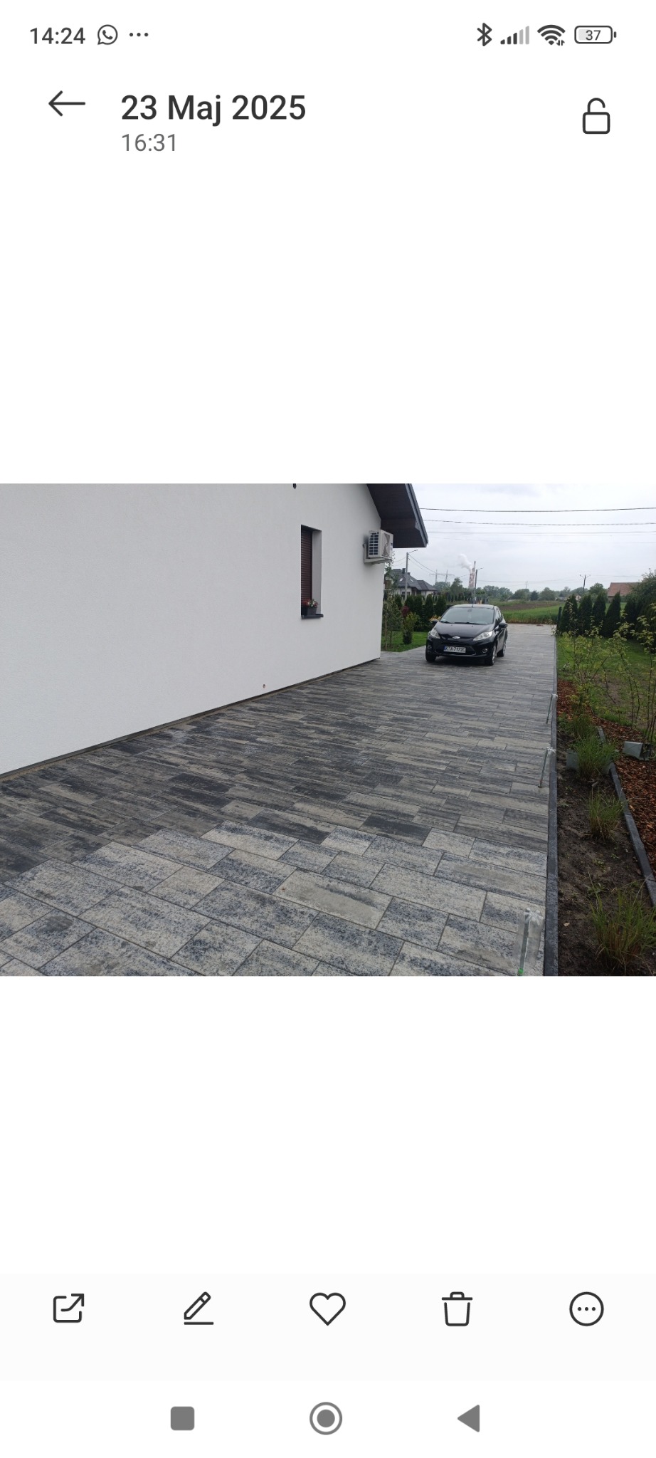 Nowoczesny dom z białą elewacją i brukowanym podjazdem z szarej kostki. Na podjeździe stoi czarny samochód osobowy. Widoczne elementy zieleni.