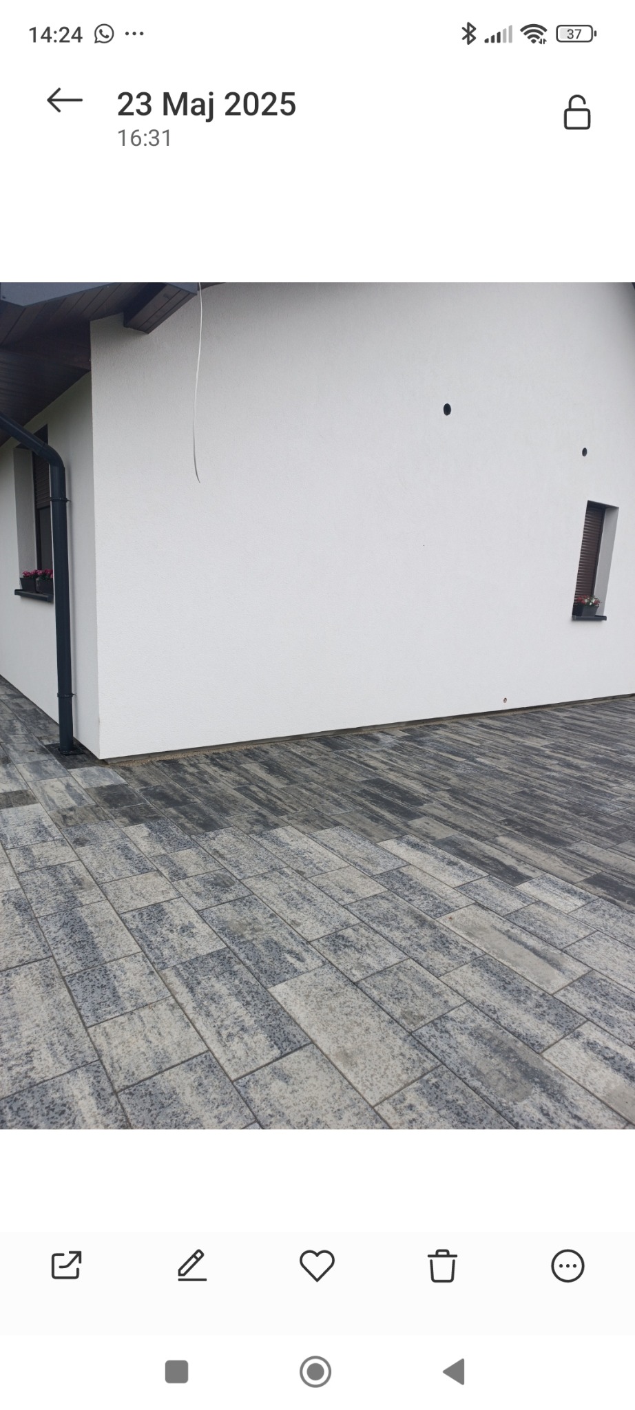 Biała elewacja domu z oknem i kwiatami, widok z poziomu kostki brukowej. Minimalistyczny design, ciemna rynna, nowoczesne wykończenie.