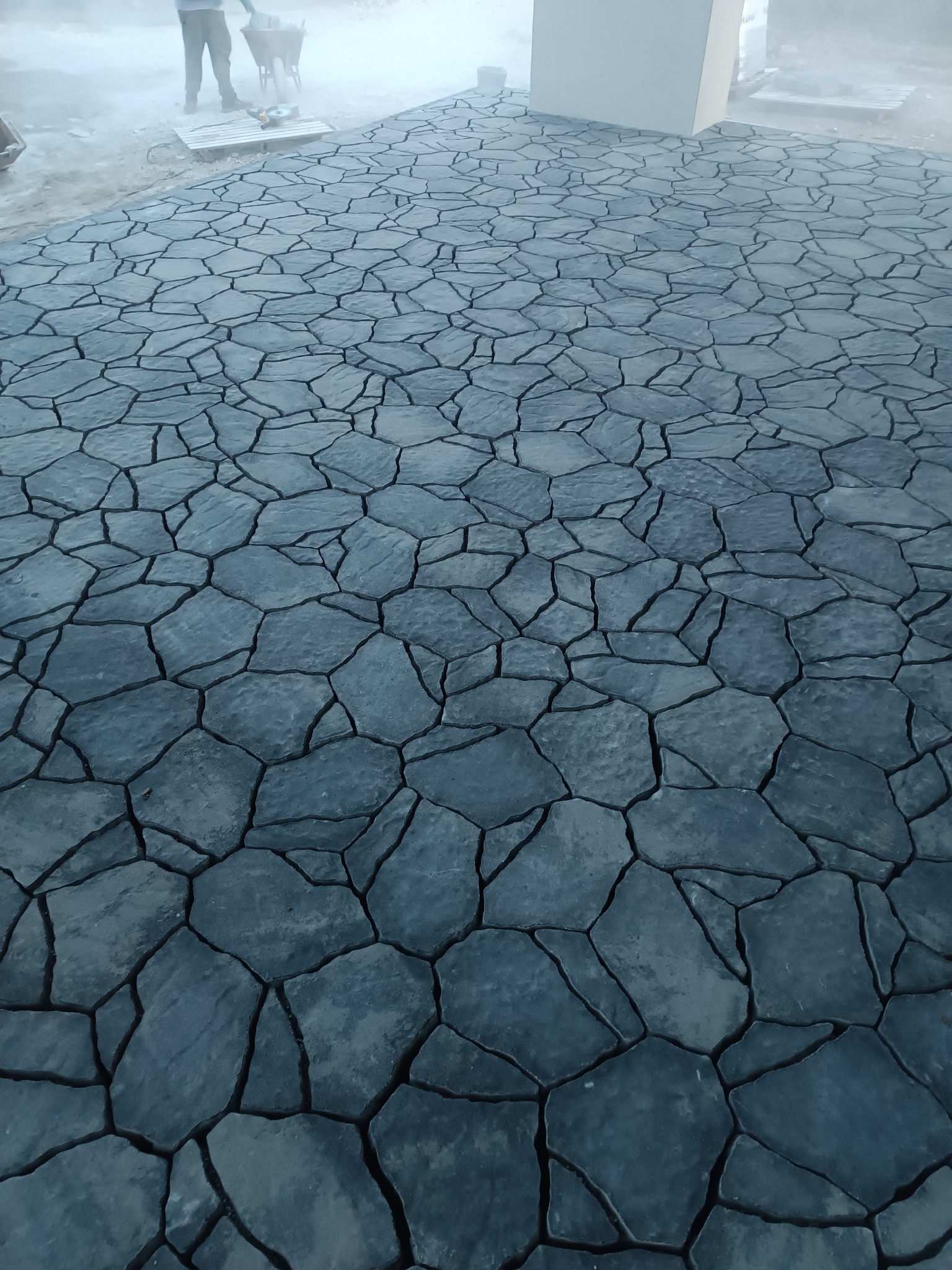 Ułożona, szara kostka brukowa o nieregularnym kształcie, imitująca kamień, tworząca wzór na podjeździe. W tle widoczna osoba z taczką i narzędzia.