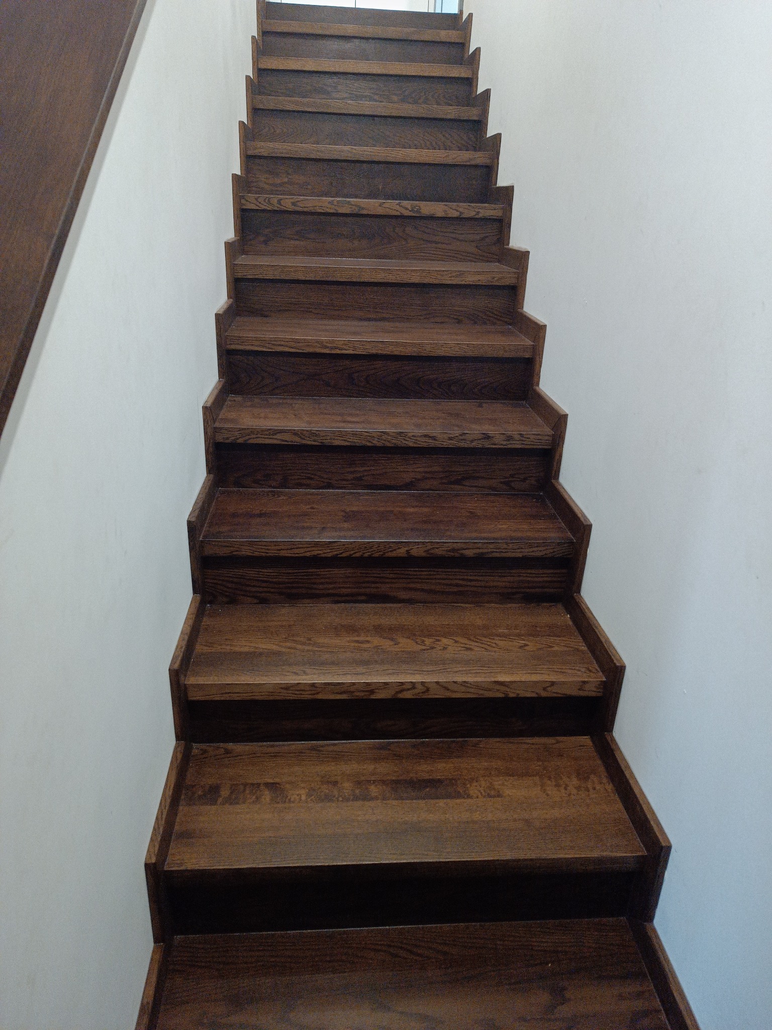 Drewniane schody w odcieniach brązu, widok z dołu, ukazujący strukturę drewna i precyzję wykonania stopni. Minimalistyczne białe ściany podkreślają ciepło drewna.