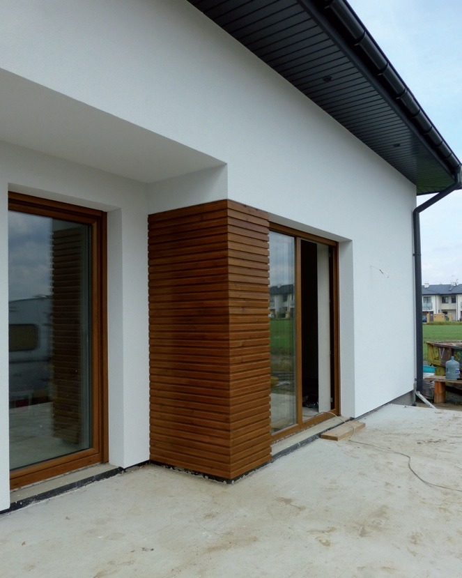 Biała elewacja domu z drewnianą okładziną narożną i oknami z brązowymi ramami. Widok z zewnątrz, betonowe podłoże.