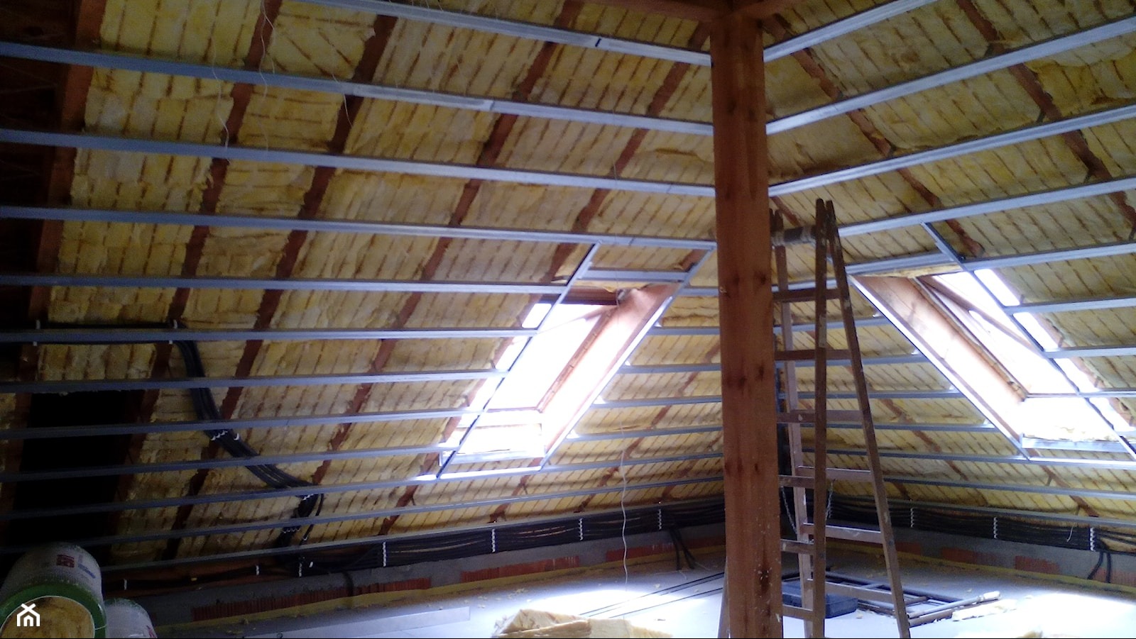 Ocieplenie poddasza wełną mineralną, widoczne metalowe profile, okno dachowe i drewniana drabina. Prace w toku, widok od dołu.