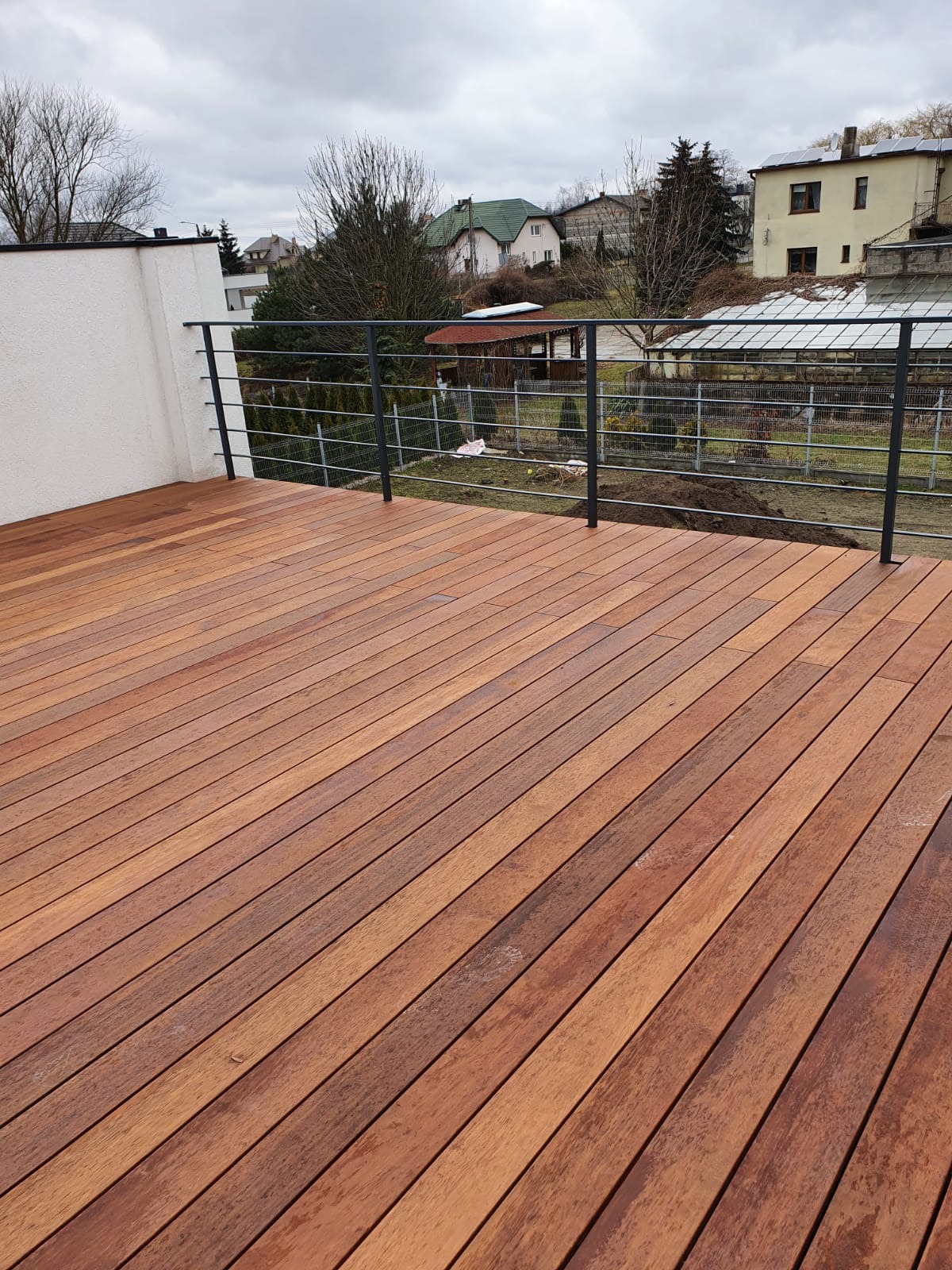 Nowoczesny taras z egzotycznego drewna, zabezpieczony metalową balustradą. Widok na ogród i zabudowę jednorodzinną w tle, pochmurne niebo.