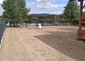 Przygotowany teren pod budowę z widokiem na jezioro i domy w tle. Piasek wyrównany, ogrodzenie po lewej, elementy konstrukcyjne po prawej.
