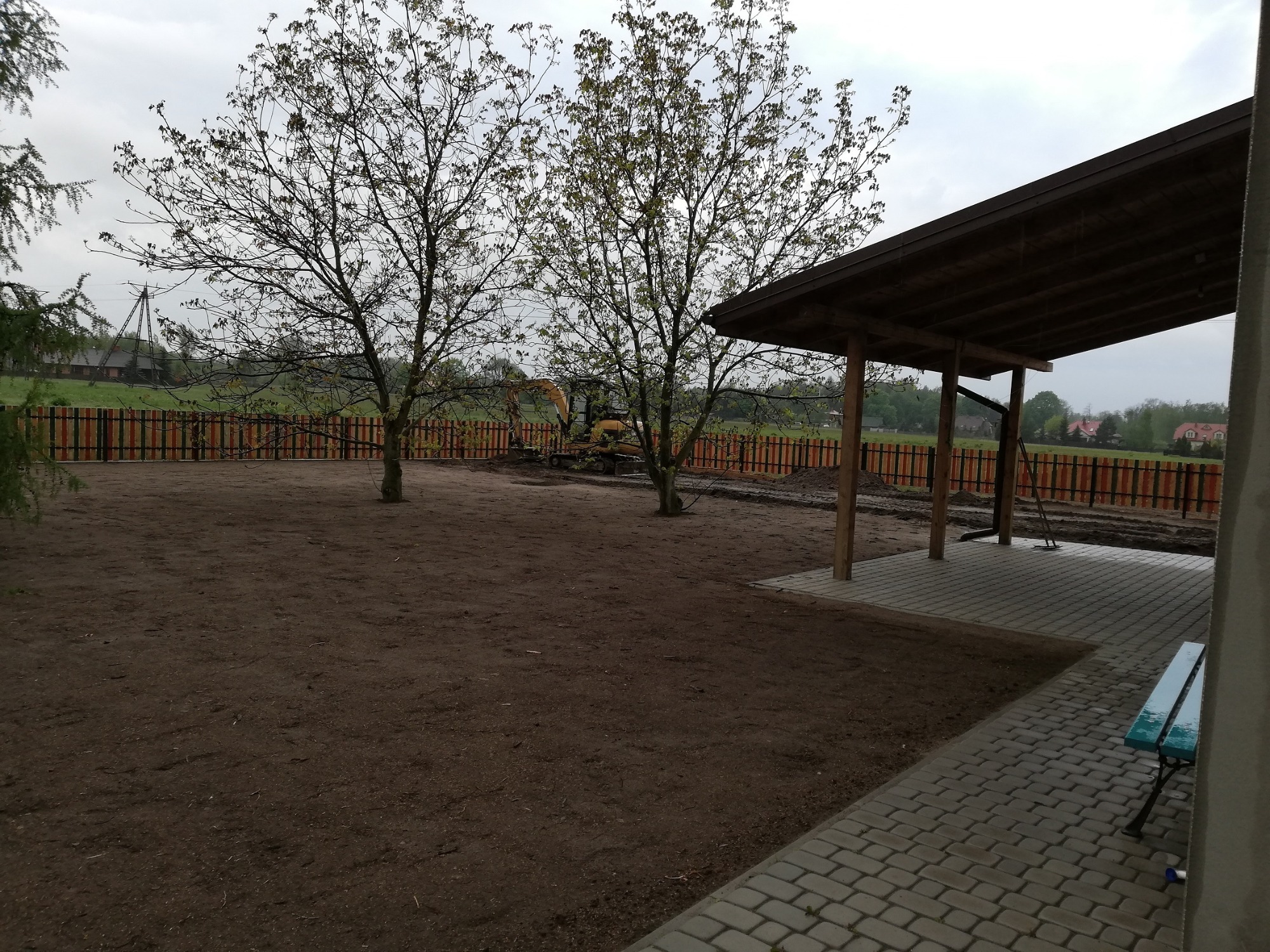 Przygotowany teren pod budowę z widoczną, kolorową, drewnianą palisadą, wykopem i zadaszeniem. W tle drzewa i pochmurne niebo.