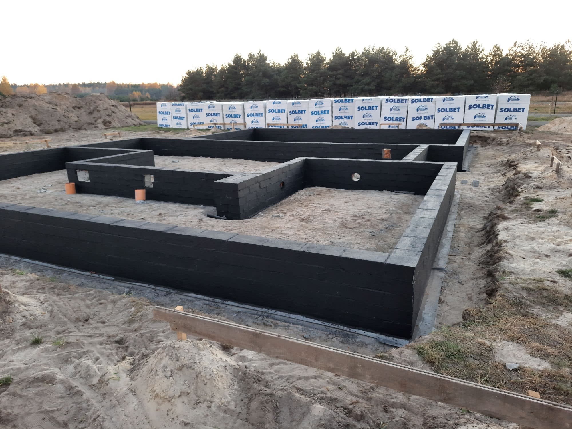 Wzniesione ściany fundamentowe z bloczków Solbet, zabezpieczone czarną izolacją, na tle piaszczystego terenu i zapasu bloczków. Widok z poziomu gruntu.