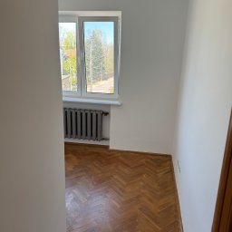 DomGardenSerwis - Wnętrze pokoju po malowaniu ścian na biało, z parkietem w jodełkę i grzejnikiem pod oknem. Widok zza framugi drzwi.