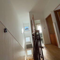 DomGardenSerwis - Jasne, odświeżone wnętrze klatki schodowej po malowaniu, z drewnianymi schodami i balustradą. Widoczne okna, drzwi i fragmenty narzędzi malarskich.
