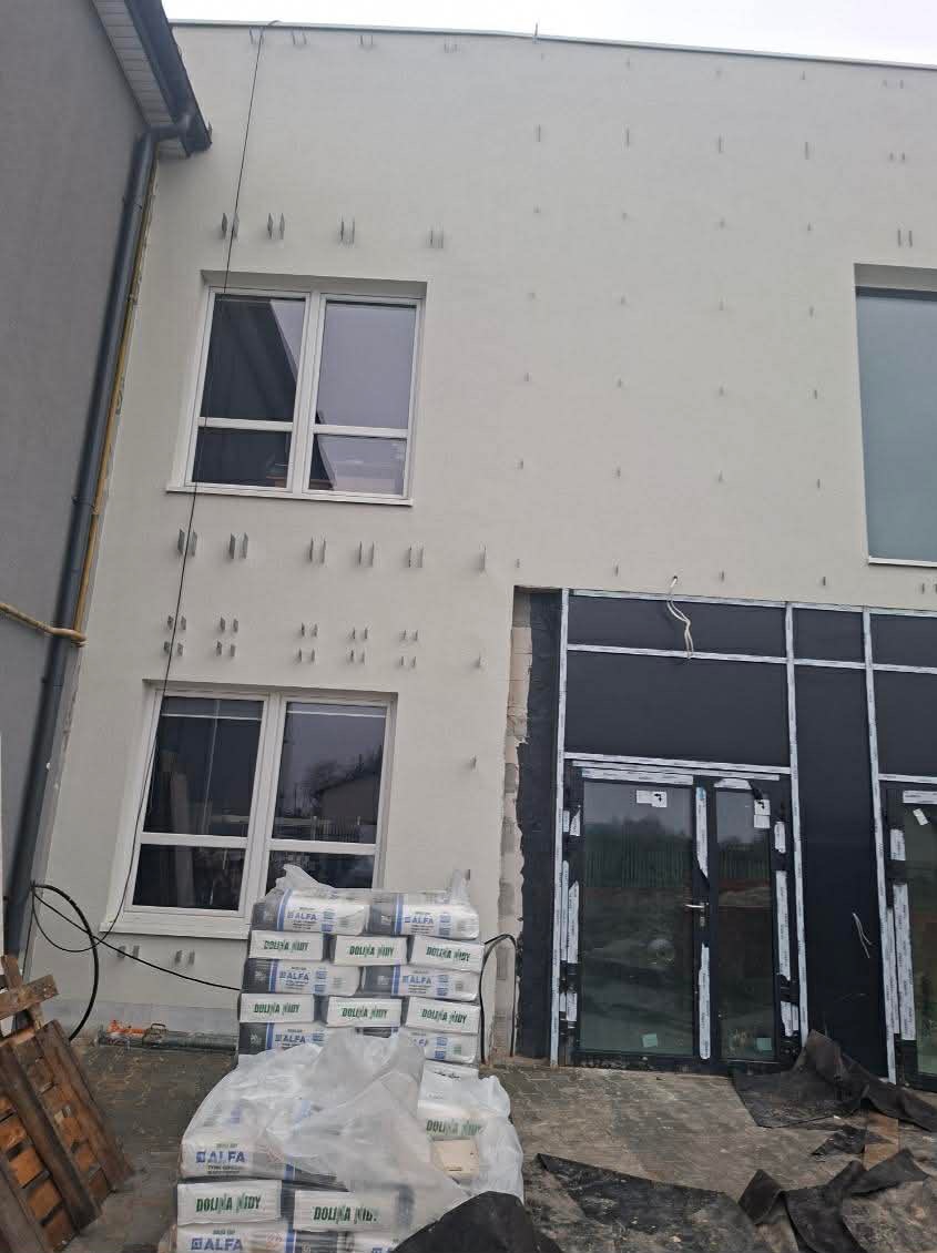 Ocieplona elewacja budynku w trakcie prac wykończeniowych. Widoczne okna, wejście z czarną stolarką i materiały izolacyjne. Mocowania na ścianie.