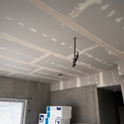 B.W.I.P. SPÓŁKA Z OGRANICZONĄ ODPOWIEDZIALNOŚCIĄ - Sufit podwieszany z płyt gipsowo-kartonowych w trakcie szpachlowania, widoczne łączenia i wkręty. W tle surowe ściany i okno z folią ochronną.