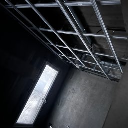 B.W.I.P. SPÓŁKA Z OGRANICZONĄ ODPOWIEDZIALNOŚCIĄ - Montaż metalowej konstrukcji sufitu podwieszanego w pomieszczeniu z oknem dachowym. Widoczne rury wentylacyjne. Surowe ściany w odcieniach szarości.