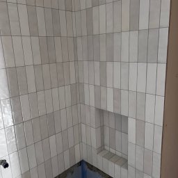 B.W.I.P. SPÓŁKA Z OGRANICZONĄ ODPOWIEDZIALNOŚCIĄ - Ułożone pionowo, podłużne, białe płytki w łazience we Wrześni. W ścianie wnęka wyłożona tymi samymi płytkami. Widoczny fragment brodzika.