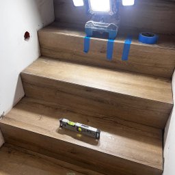 B.W.I.P. SPÓŁKA Z OGRANICZONĄ ODPOWIEDZIALNOŚCIĄ - Montaż paneli na schodach z użyciem poziomicy i taśmy malarskiej, oświetlonych lampą roboczą. Widoczne niedoskonałości ściany i otwór po gniazdku.