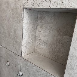 B.W.I.P. SPÓŁKA Z OGRANICZONĄ ODPOWIEDZIALNOŚCIĄ - Ściana wyłożona szarymi płytkami z wnęką. Widoczne elementy poziomowania płytek. Minimalistyczny design, surowy betonowy wygląd płytek.