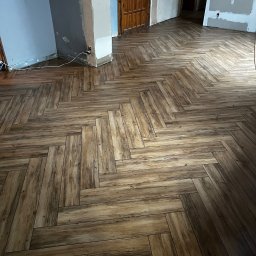 B.W.I.P. SPÓŁKA Z OGRANICZONĄ ODPOWIEDZIALNOŚCIĄ - Ukończona podłoga z paneli ułożonych w jodełkę, w odcieniach brązu, w trakcie remontu. Widoczne niedokończone ściany i futryny drzwi.