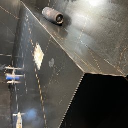 B.W.I.P. SPÓŁKA Z OGRANICZONĄ ODPOWIEDZIALNOŚCIĄ - Ciemna, błyszcząca glazura z złotymi żyłkami w kabinie prysznicowej. Widoczne elementy montażowe i głośnik JBL. Ukończone wykończenie z dbałością o detale.