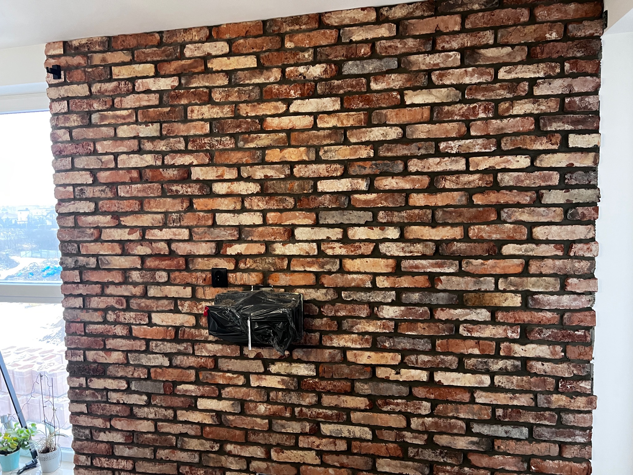 Ściana z dekoracyjnej cegły w odcieniach czerwieni i brązu, z widocznym gniazdkiem elektrycznym i elementem zabezpieczonym folią. Fragment okna z lewej strony.