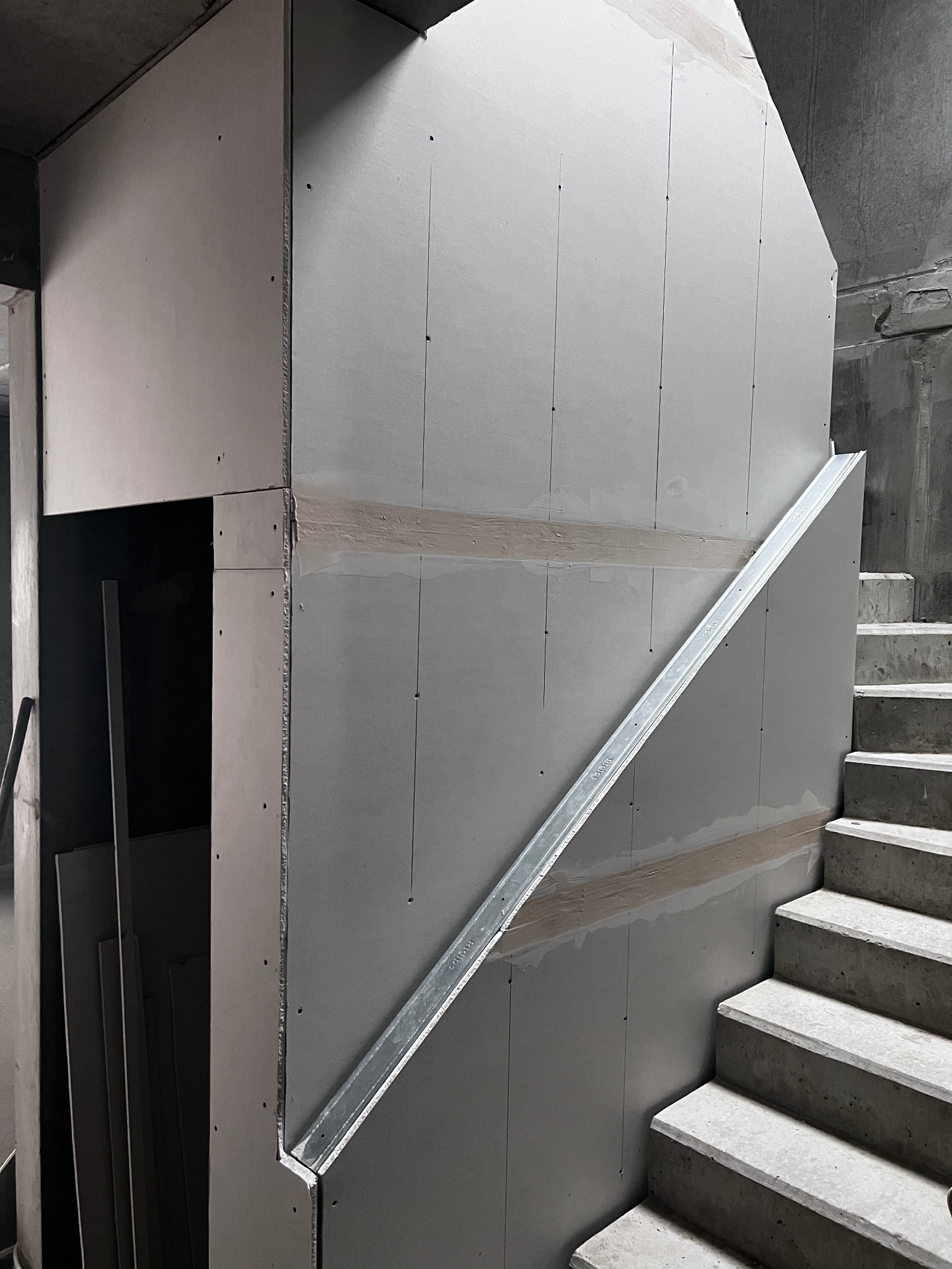 Montaż płyt gipsowo-kartonowych na ścianie przy schodach, widoczne profile i łączenia. Surowy stan wykończenia, szare odcienie betonu i gipsu.