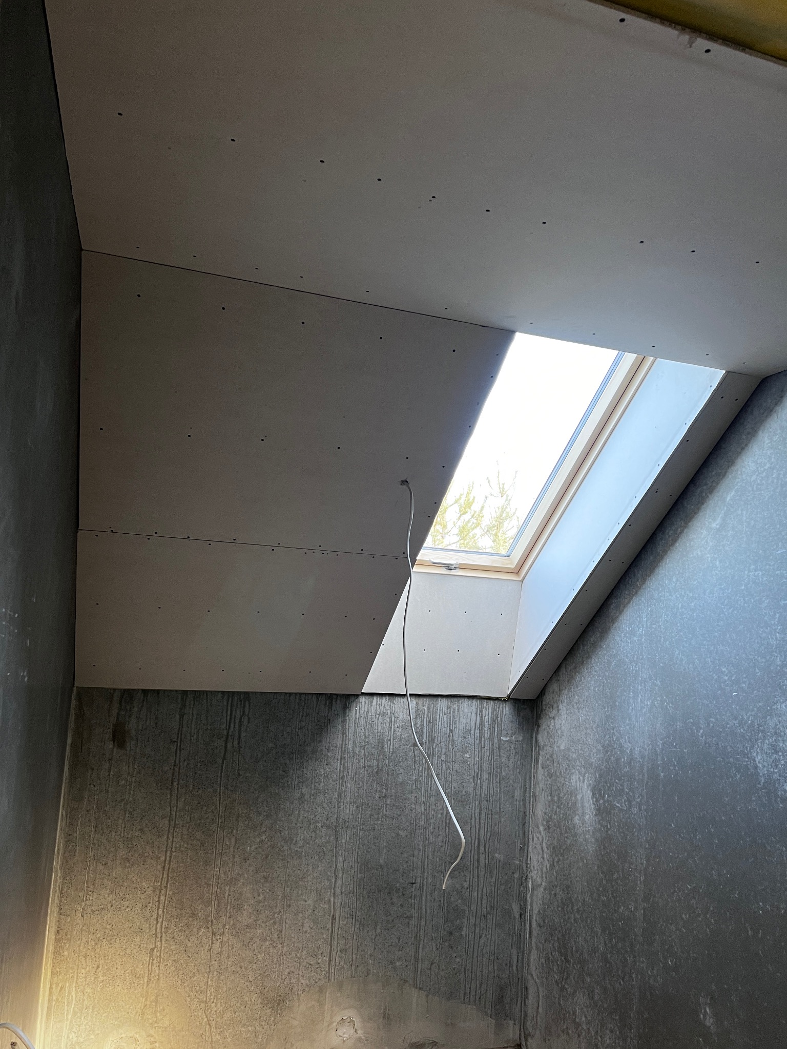 Montaż płyt gipsowo-kartonowych na poddaszu z oknem dachowym. Widoczne łączenia płyt i surowe ściany. Kabel zwisa z sufitu. Etap prac wykończeniowych.