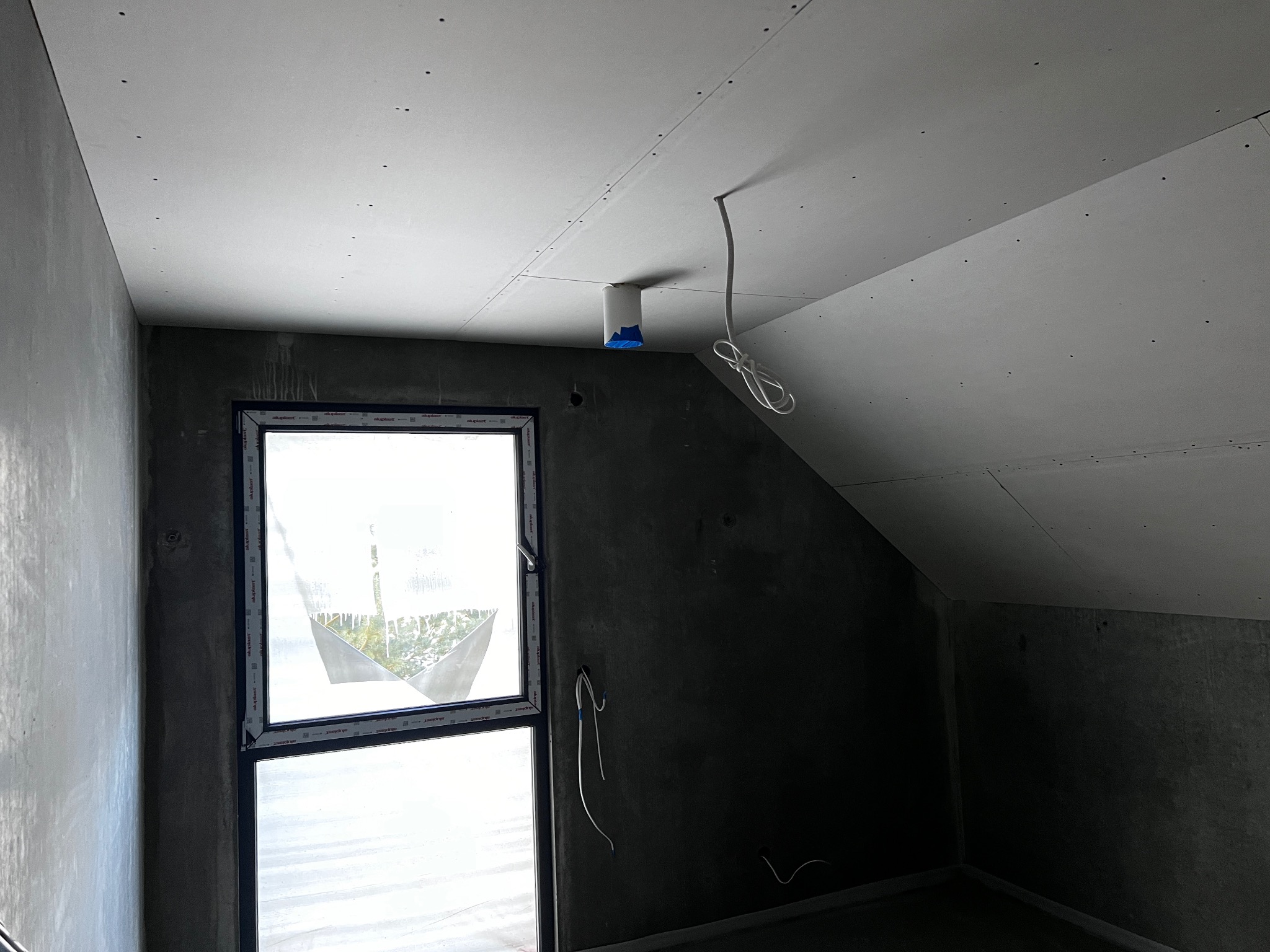 Poddasze w trakcie prac wykończeniowych: zamontowane płyty gipsowo-kartonowe na skosach sufitu, surowe ściany, okno z folią ochronną. Widoczne przewody elektryczne.