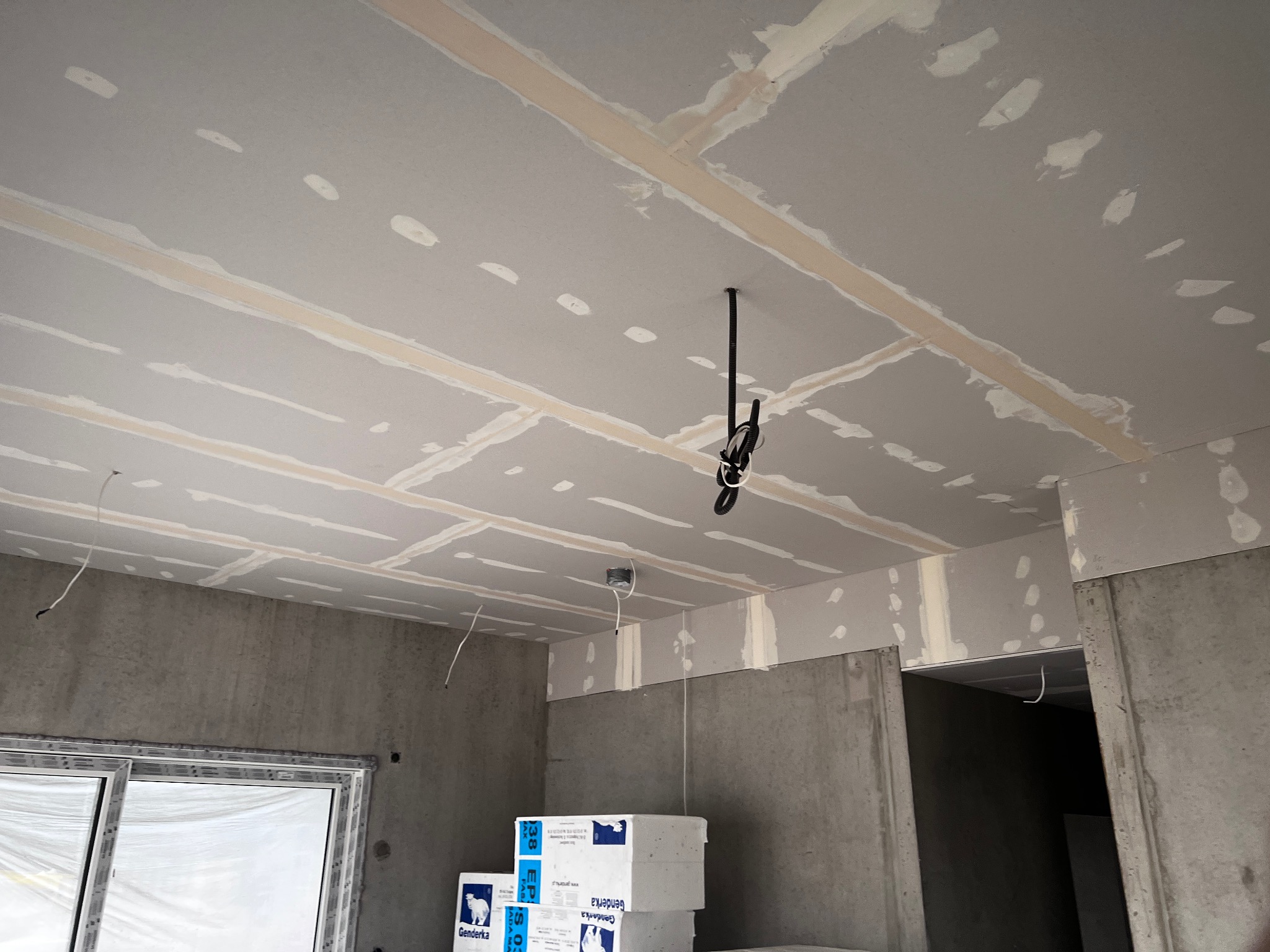 Sufit podwieszany z płyt gipsowo-kartonowych w trakcie szpachlowania, widoczne łączenia i wkręty. W tle surowe ściany i okno z folią ochronną.