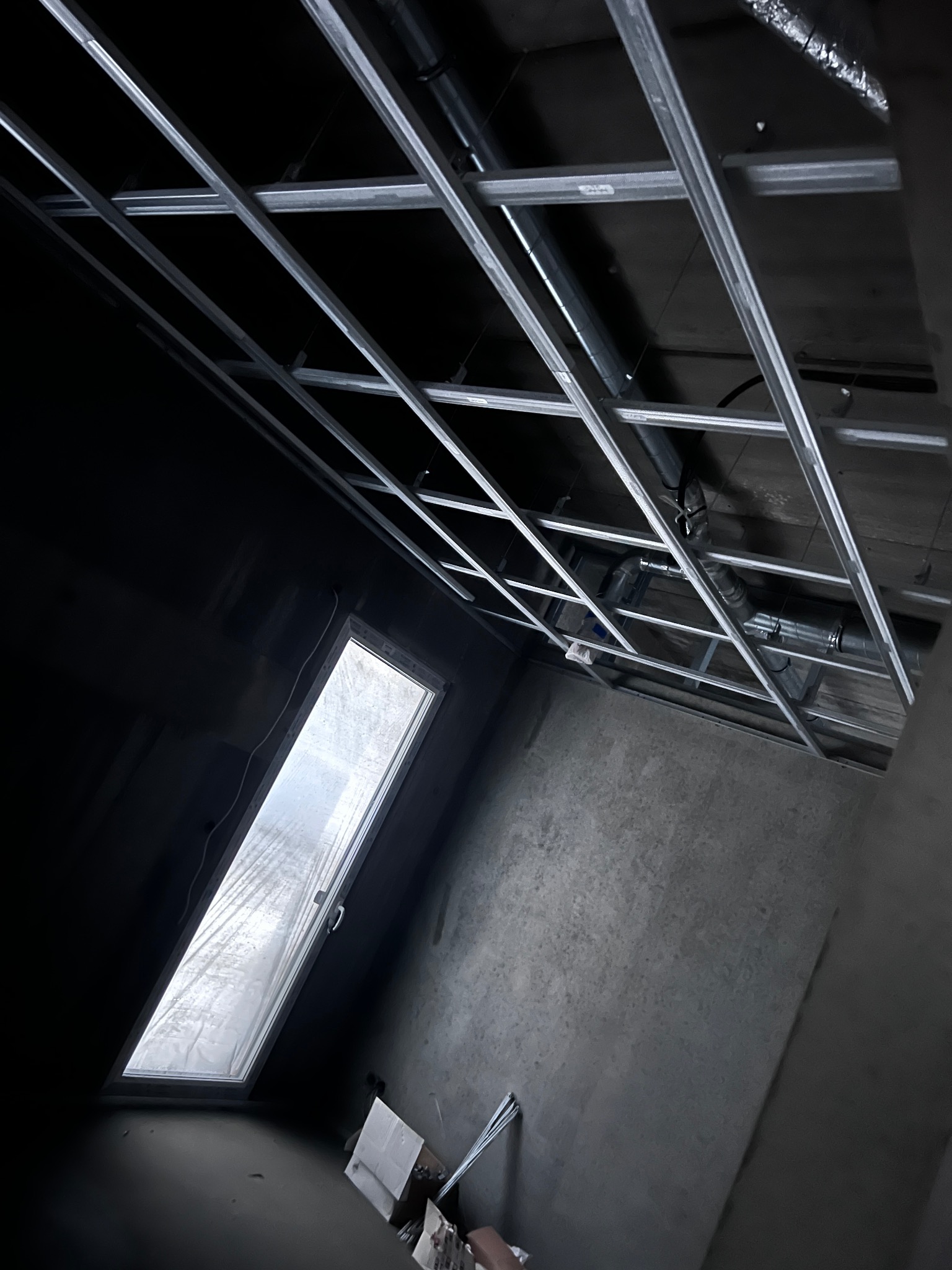 Montaż metalowej konstrukcji sufitu podwieszanego w pomieszczeniu z oknem dachowym. Widoczne rury wentylacyjne. Surowe ściany w odcieniach szarości.