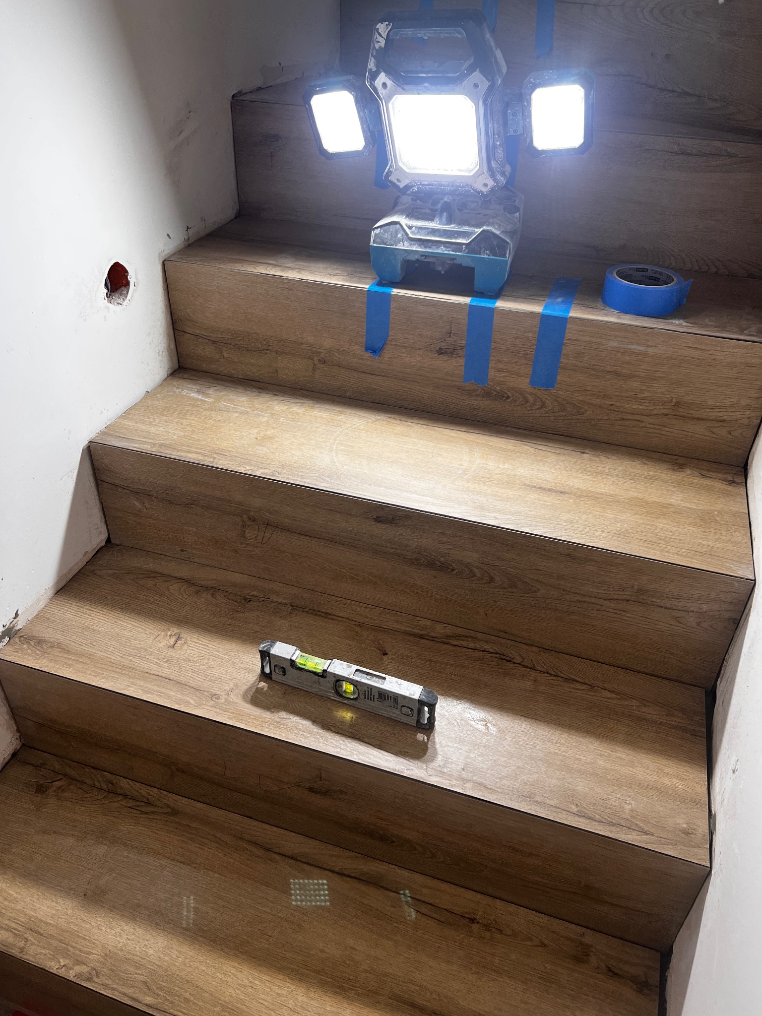 Montaż paneli na schodach z użyciem poziomicy i taśmy malarskiej, oświetlonych lampą roboczą. Widoczne niedoskonałości ściany i otwór po gniazdku.