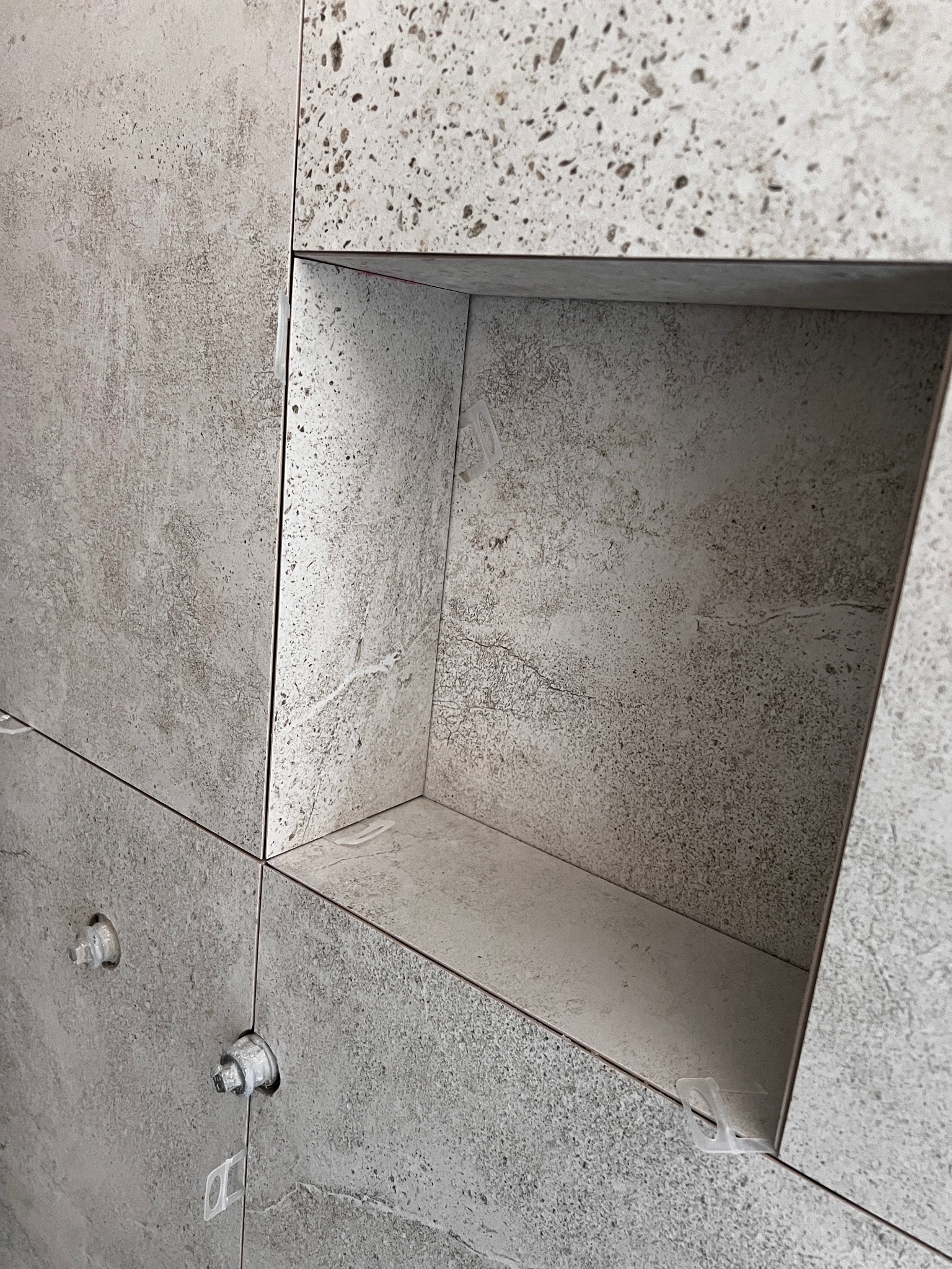 Ściana wyłożona szarymi płytkami z wnęką. Widoczne elementy poziomowania płytek. Minimalistyczny design, surowy betonowy wygląd płytek.