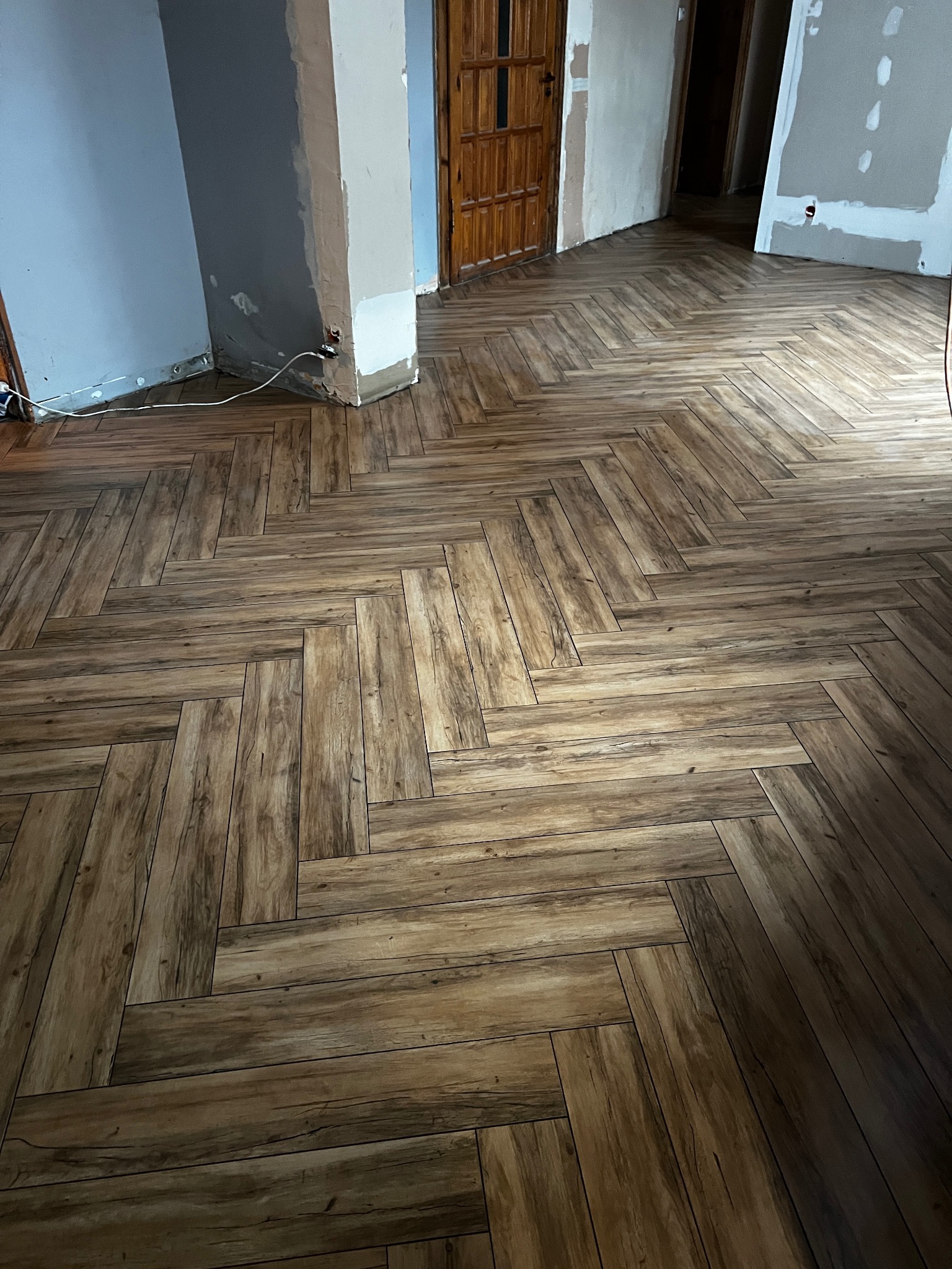 Ukończona podłoga z paneli ułożonych w jodełkę, w odcieniach brązu, w trakcie remontu. Widoczne niedokończone ściany i futryny drzwi.