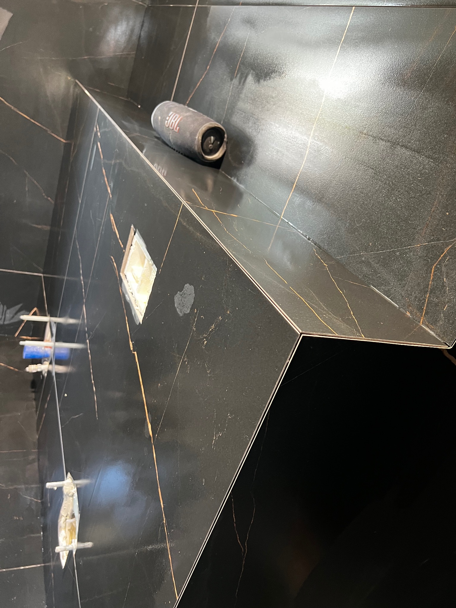 Ciemna, błyszcząca glazura z złotymi żyłkami w kabinie prysznicowej. Widoczne elementy montażowe i głośnik JBL. Ukończone wykończenie z dbałością o detale.