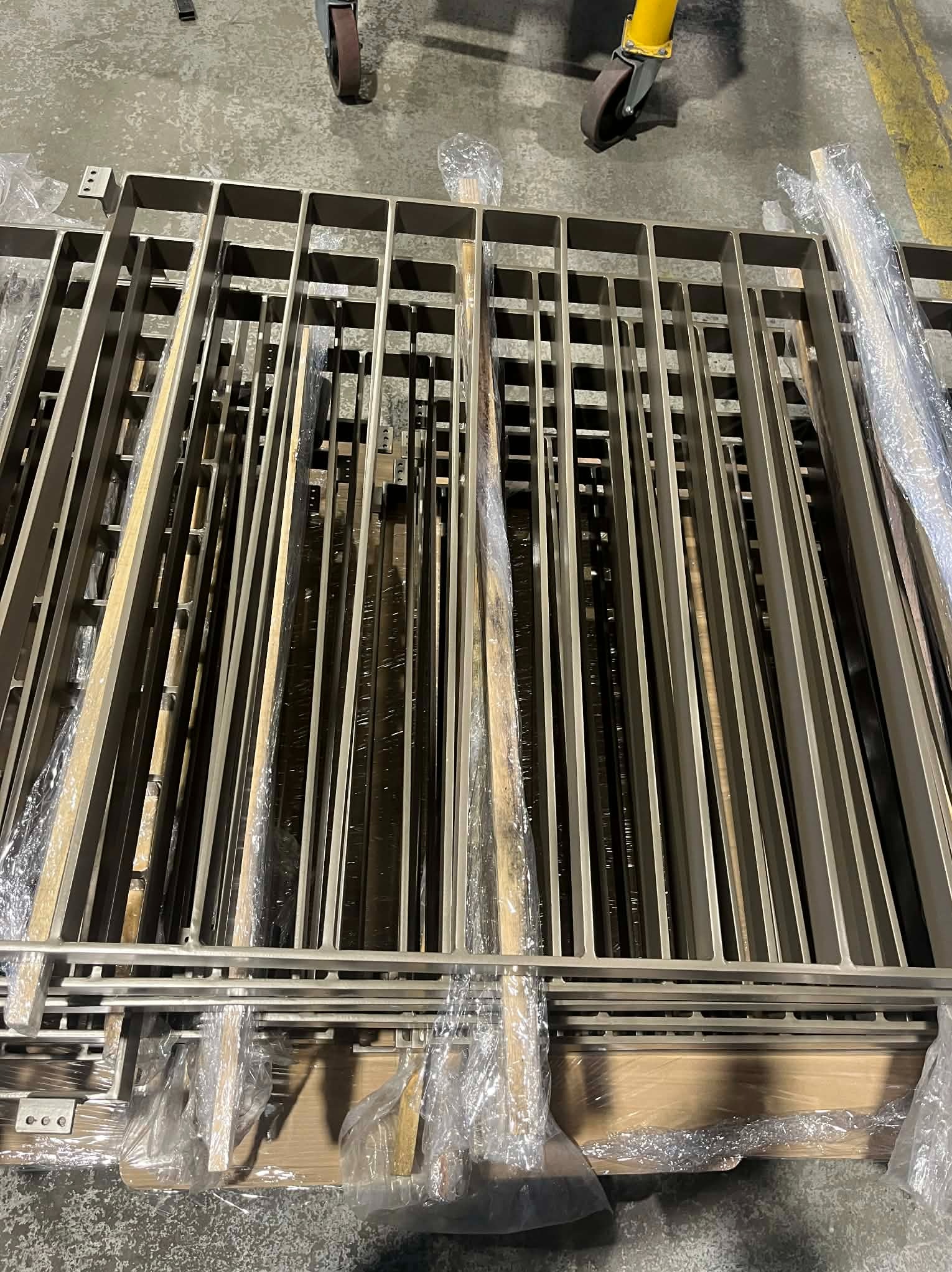 Stos metalowych, spawanych elementów konstrukcyjnych, prawdopodobnie balustrad lub ogrodzeń, zabezpieczonych folią, leżących na podłodze hali produkcyjnej.