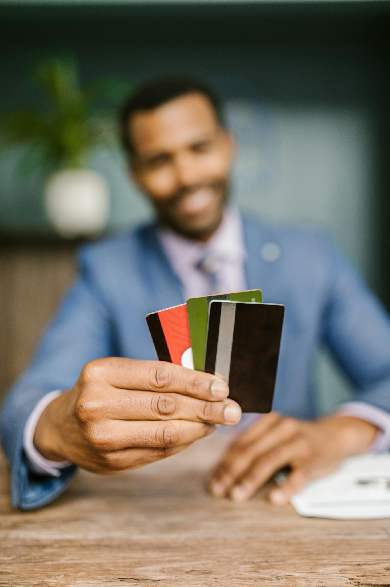 Uśmiechnięty mężczyzna w garniturze prezentuje wachlarz kart kredytowych w dłoni, siedząc przy drewnianym stole. W tle widoczna roślina doniczkowa.