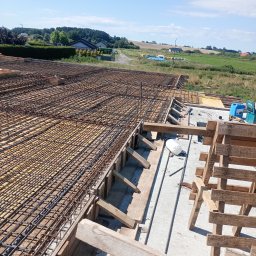 Szymon Makowiecki - Zbrojenie stropu domu jednorodzinnego z widocznymi elementami szalunku i narzędziami w tle krajobrazu wiejskiego pod błękitnym niebem.