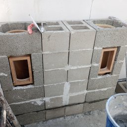 Szymon Makowiecki - Wznoszenie konstrukcji z szarych bloczków betonowych z otworami, widoczne narzędzia murarskie i poziomica. Budowa kominka lub grilla w trakcie realizacji.