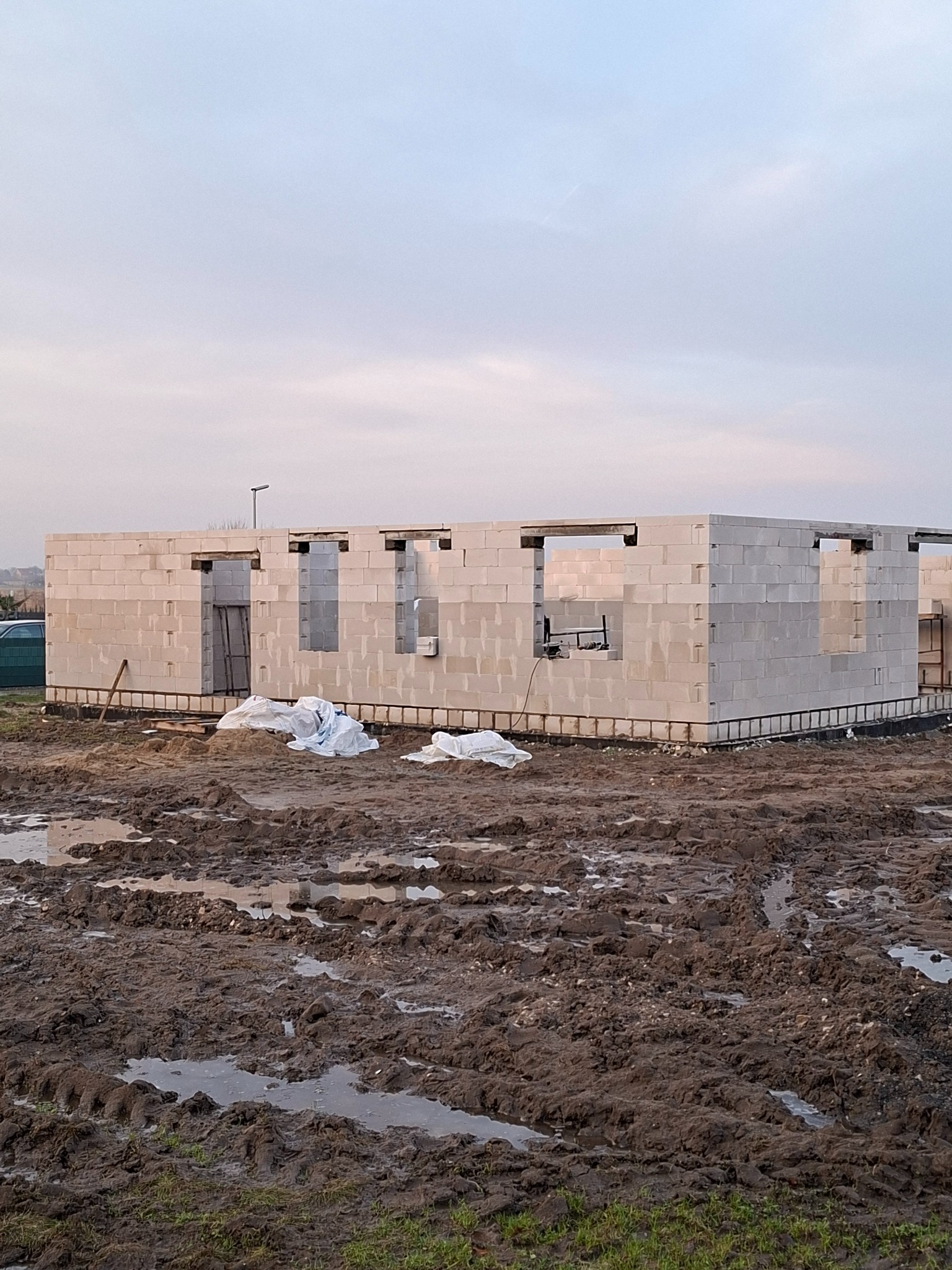 Wznoszenie ścian parterowego domu z betonu komórkowego. Widoczne otwory okienne i drzwiowe, teren budowy z błotem. Częściowo ukończony etap budowy.