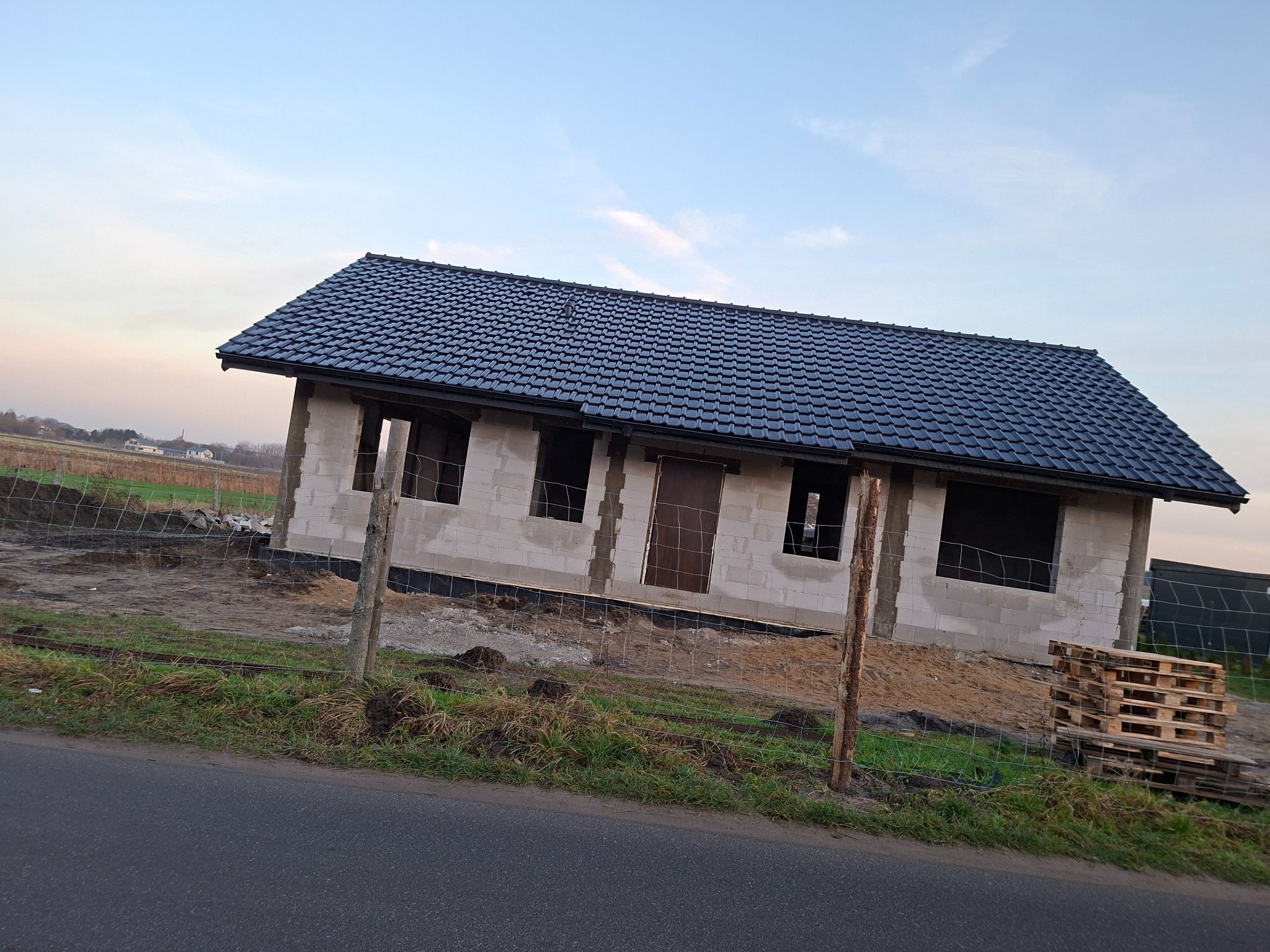 Surowy dom z ciemną dachówką, widoczny zza ogrodzenia. Ściany z betonu komórkowego, otwory okienne i drzwiowe. Palety obok. Krajobraz wiejski w tle.