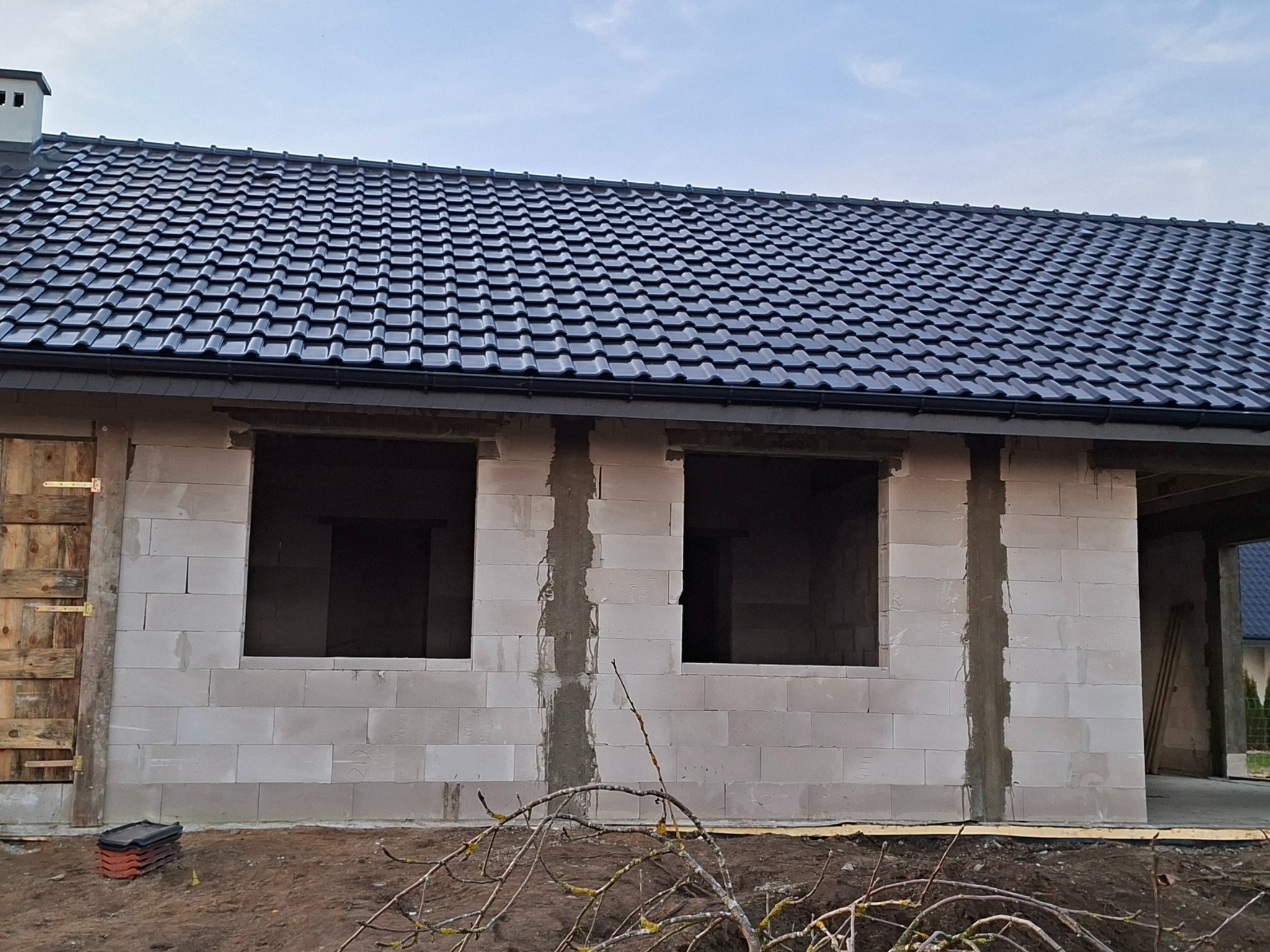 Surowy stan domu z białych bloczków, z czarnym dachem z dachówki. Widoczne otwory okienne i prowizoryczne drzwi. Etap budowy przed ociepleniem.