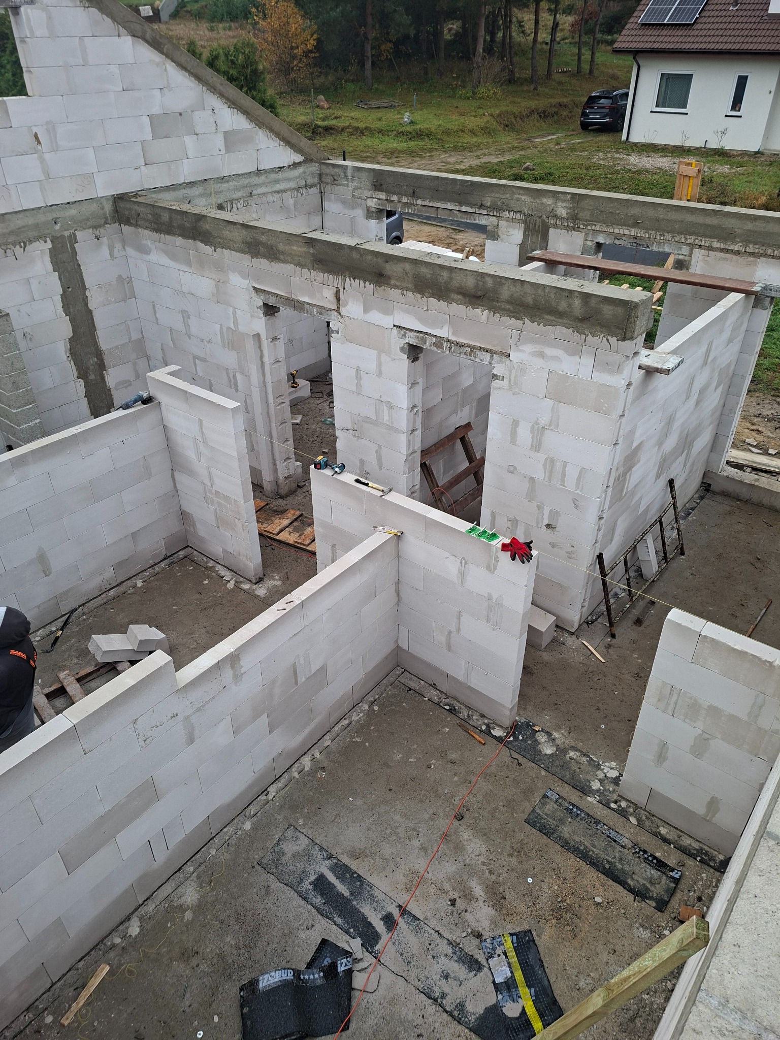 Wnętrze domu w trakcie budowy: białe ściany z betonu komórkowego, widoczne narzędzia i drabina. Na zewnątrz dom z panelami słonecznymi i samochód.