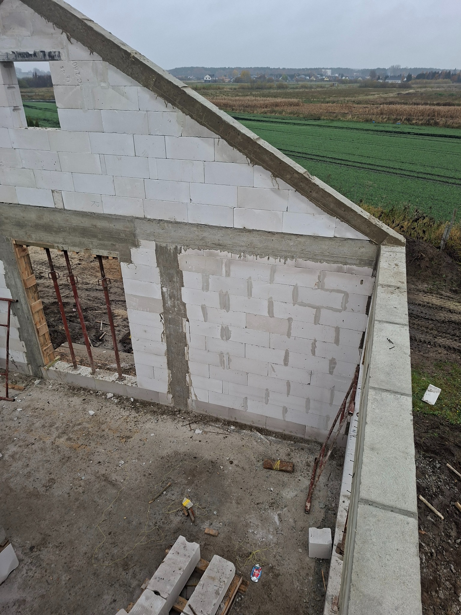 Wznoszenie ścian z betonu komórkowego na budowie domu jednorodzinnego. Widoczne podpory i narzędzia murarskie. W tle krajobraz polny pod pochmurnym niebem.