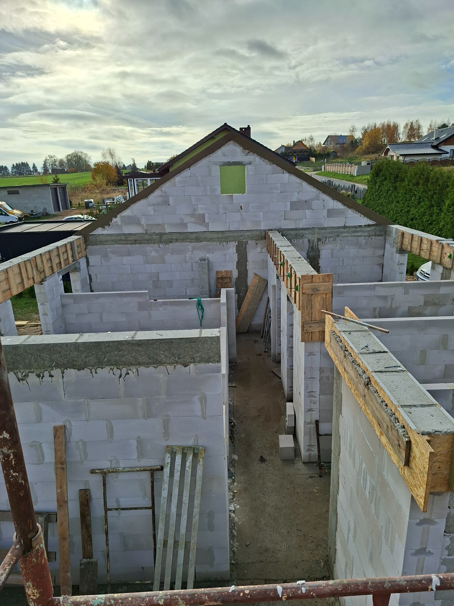 Wysoki kąt na dom w trakcie budowy z białych bloków, widoczne drewniane szalunki i zbrojenia, w tle zabudowa jednorodzinna i pochmurne niebo.