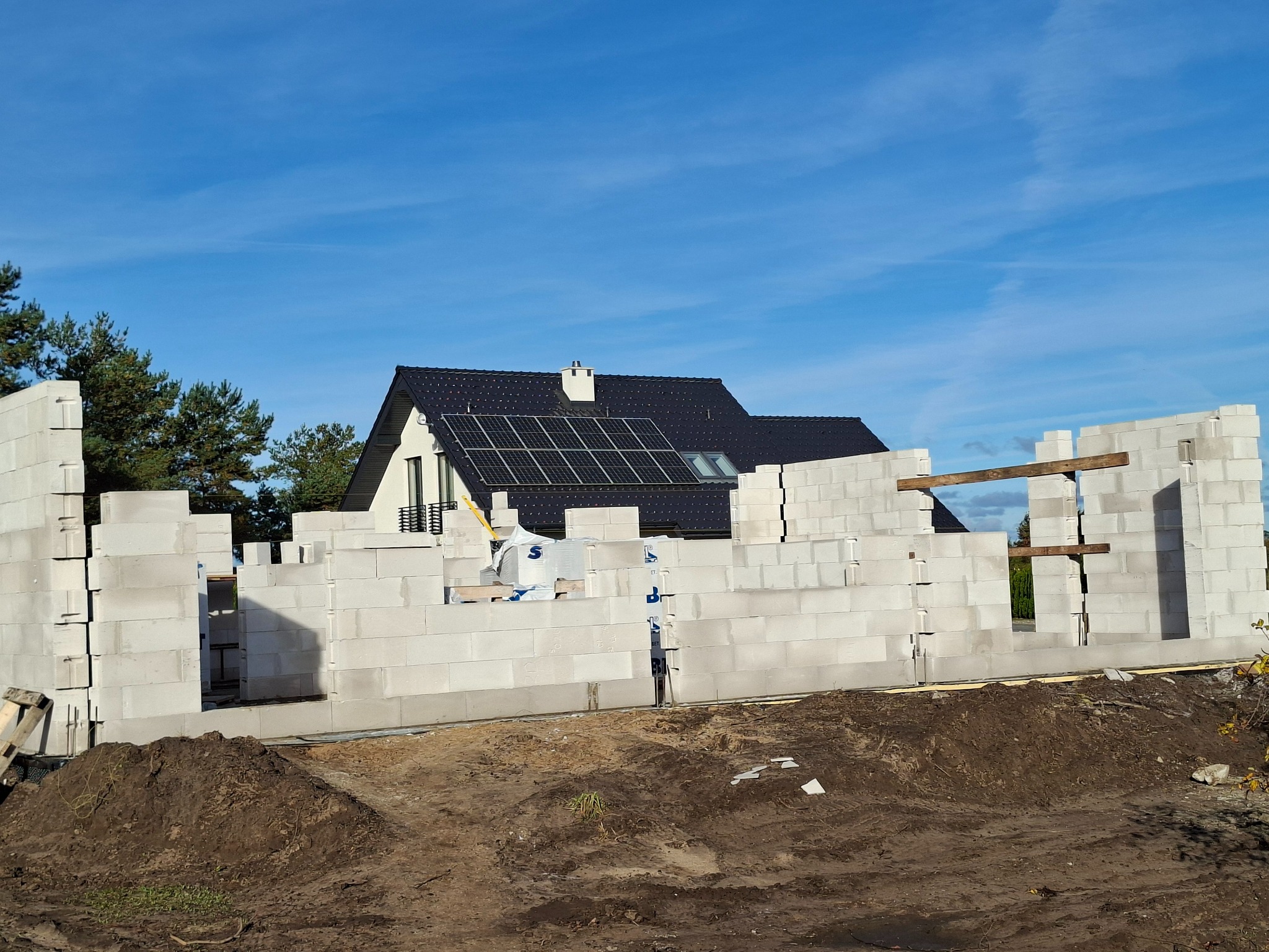 Wznoszenie ścian domu z białych bloczków betonu komórkowego. Widoczne drewniane nadproża okienne i dach z panelami solarnymi w tle, na tle błękitnego nieba.