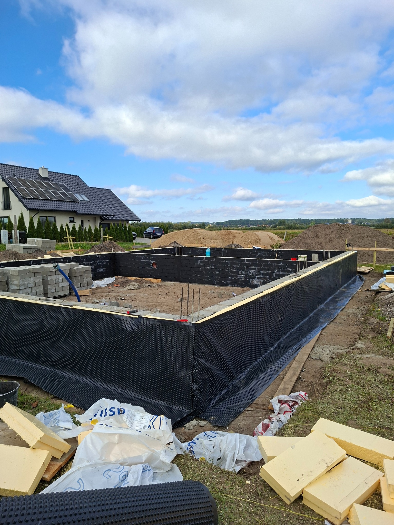 Fundamenty domu w trakcie budowy, zabezpieczone czarną folią kubełkową, z widocznymi bloczkami i izolacją termiczną. W tle dom z panelami słonecznymi i błękitne niebo.