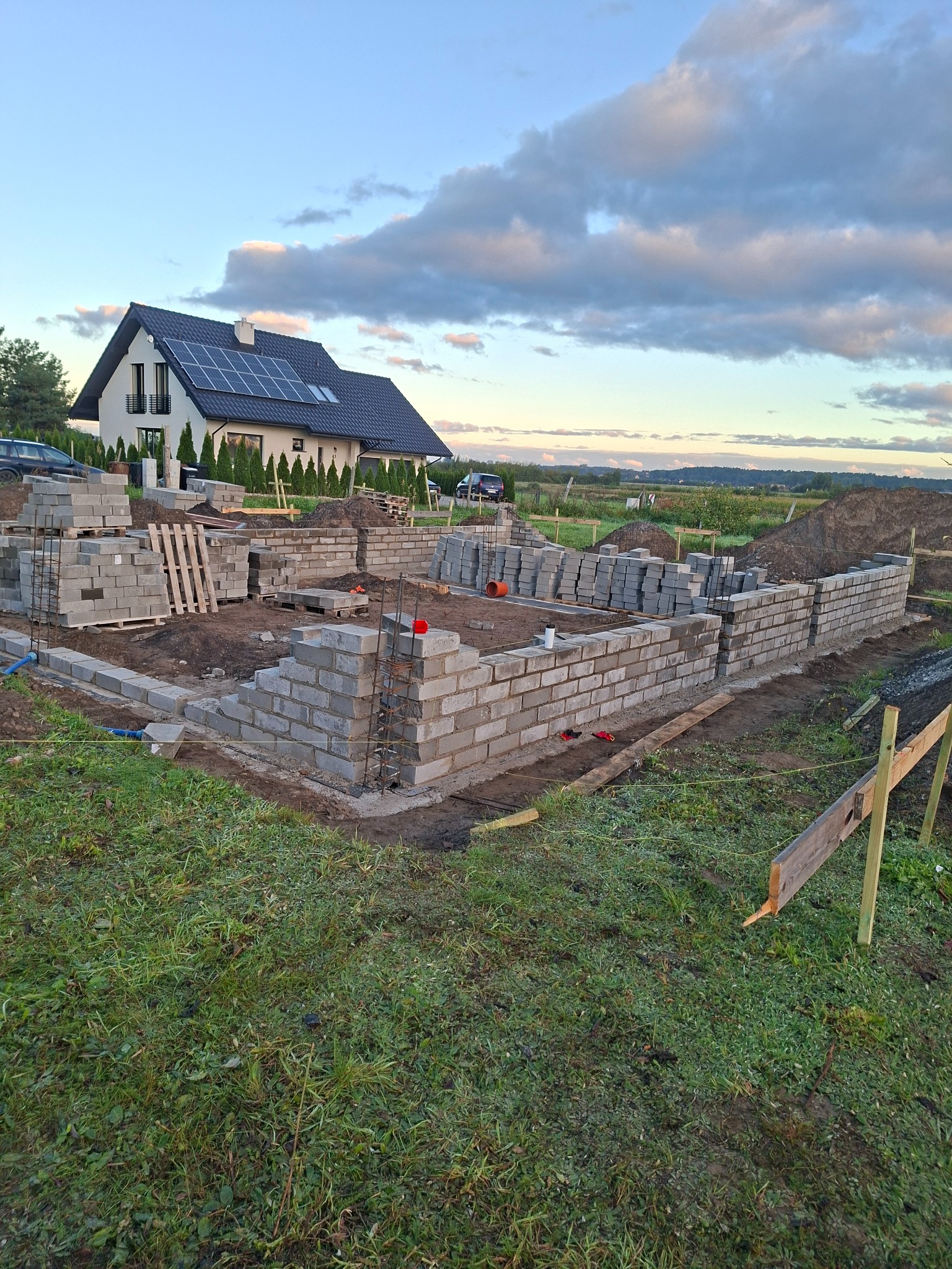 Wznoszenie fundamentów domu z bloczków betonowych, widoczne zbrojenie i przygotowane materiały. W tle dom jednorodzinny z panelami słonecznymi i błękitne niebo.