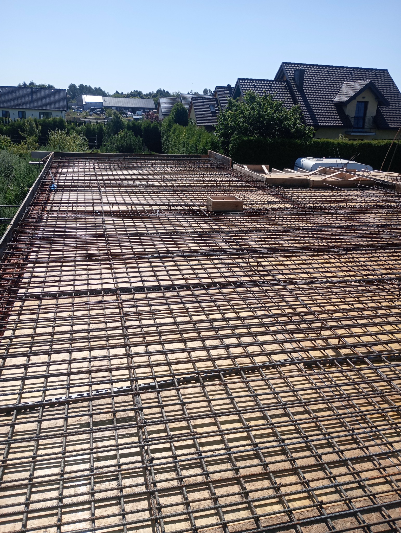 Zbrojenie stropu na deskowaniu, widok z góry. Stalowe pręty tworzą regularną siatkę. W tle zabudowa jednorodzinna i błękitne niebo. Jasne oświetlenie.