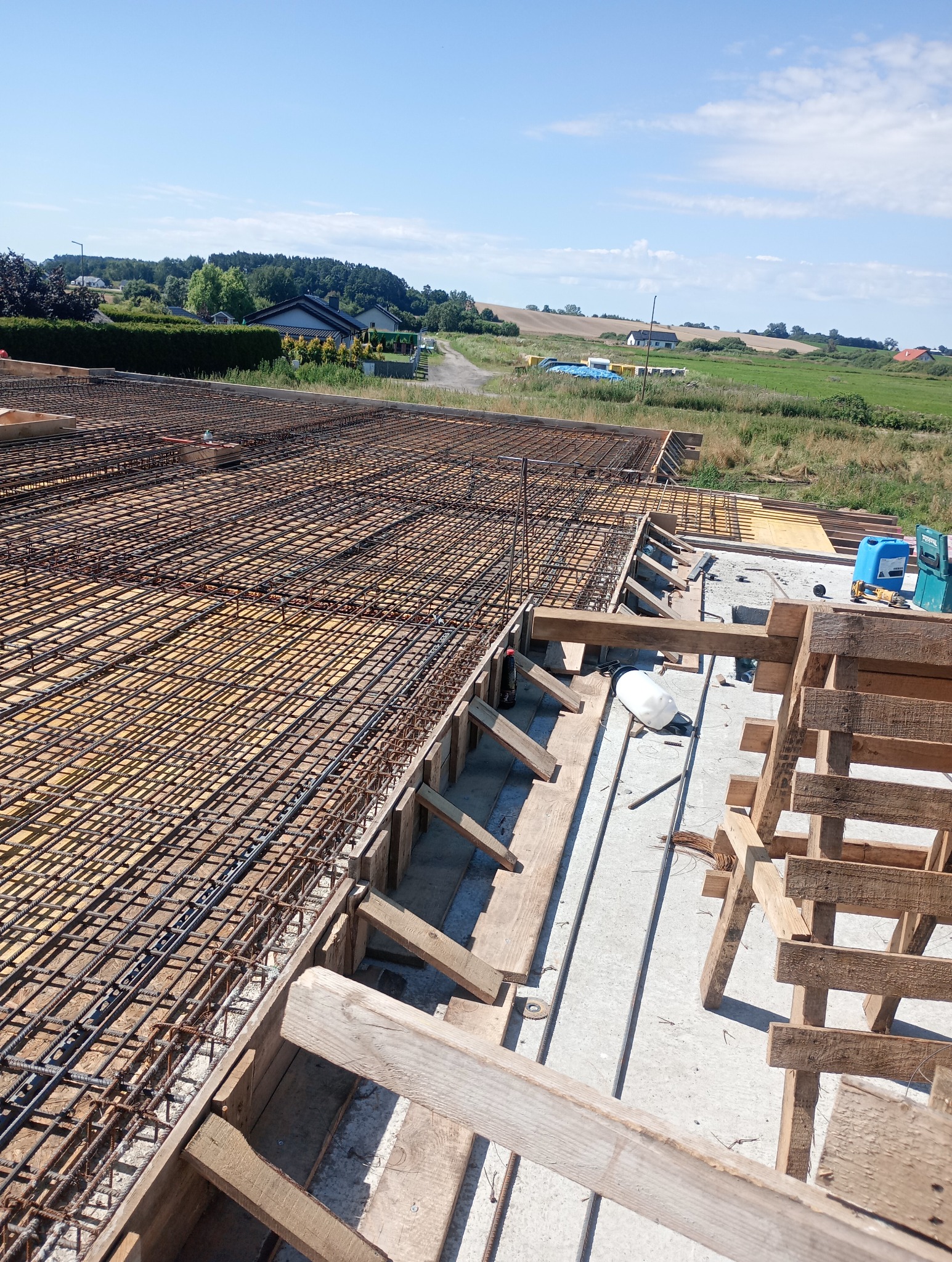 Zbrojenie stropu domu jednorodzinnego z widocznymi elementami szalunku i narzędziami w tle krajobrazu wiejskiego pod błękitnym niebem.