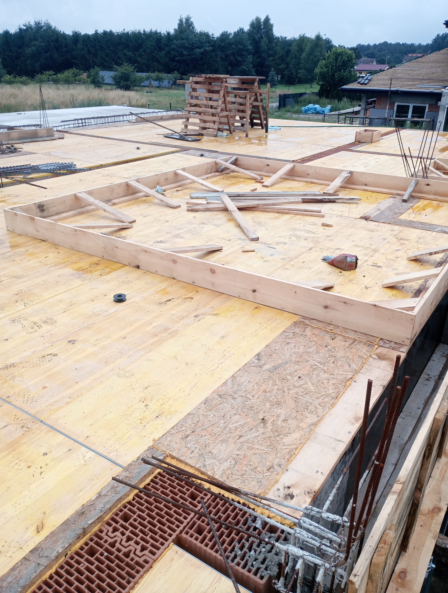 Szczegółowy widok z góry na drewniany szalunek fundamentów z ceglanymi elementami i wystającymi prętami zbrojeniowymi, przygotowany do zalania betonem.