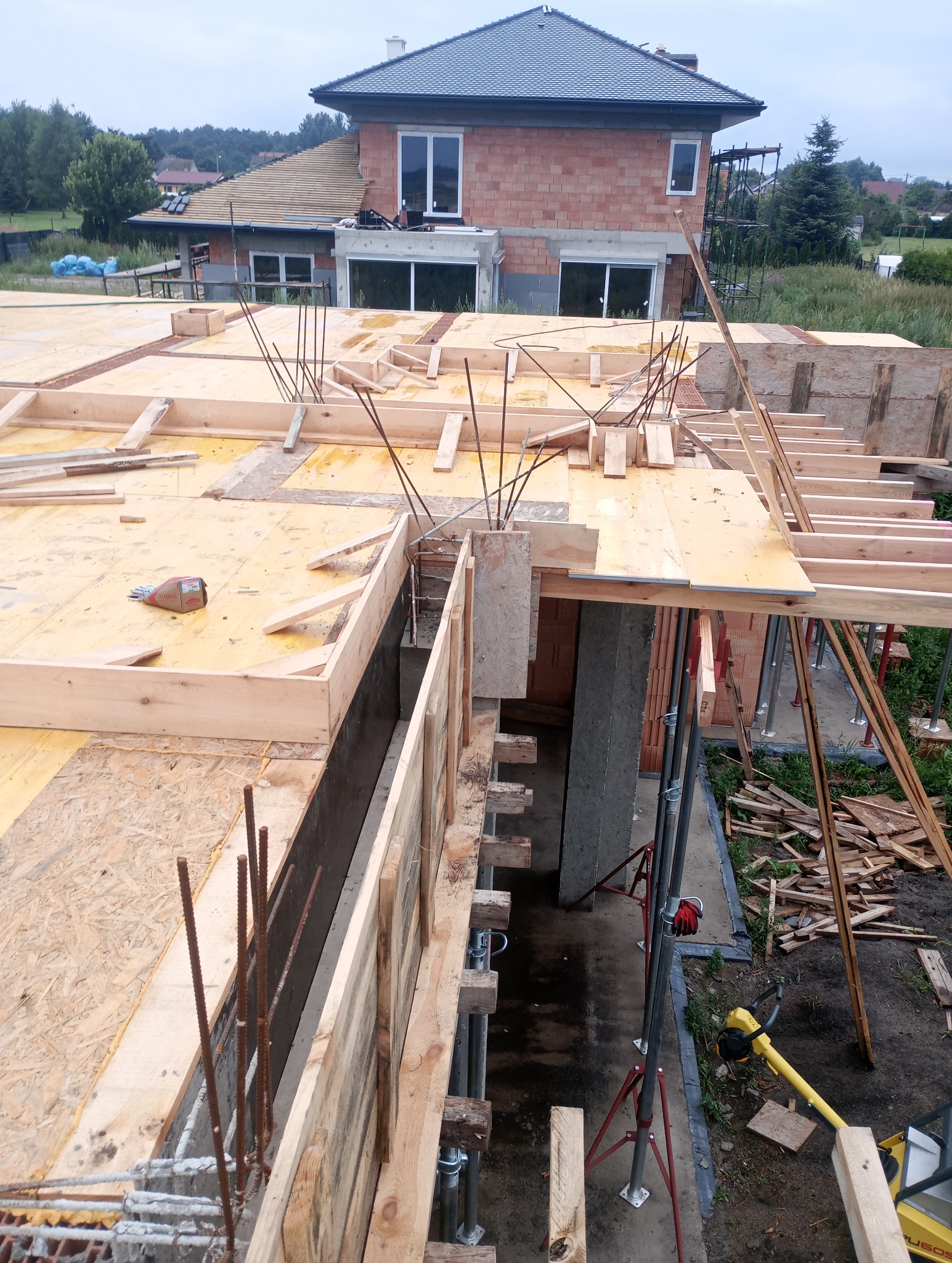 Budowa domu w trakcie: widok z góry na szalunek stropu z wystającymi prętami zbrojeniowymi. W tle wzniesione mury z czerwonej cegły. Widoczne narzędzia i materiały budowlane.