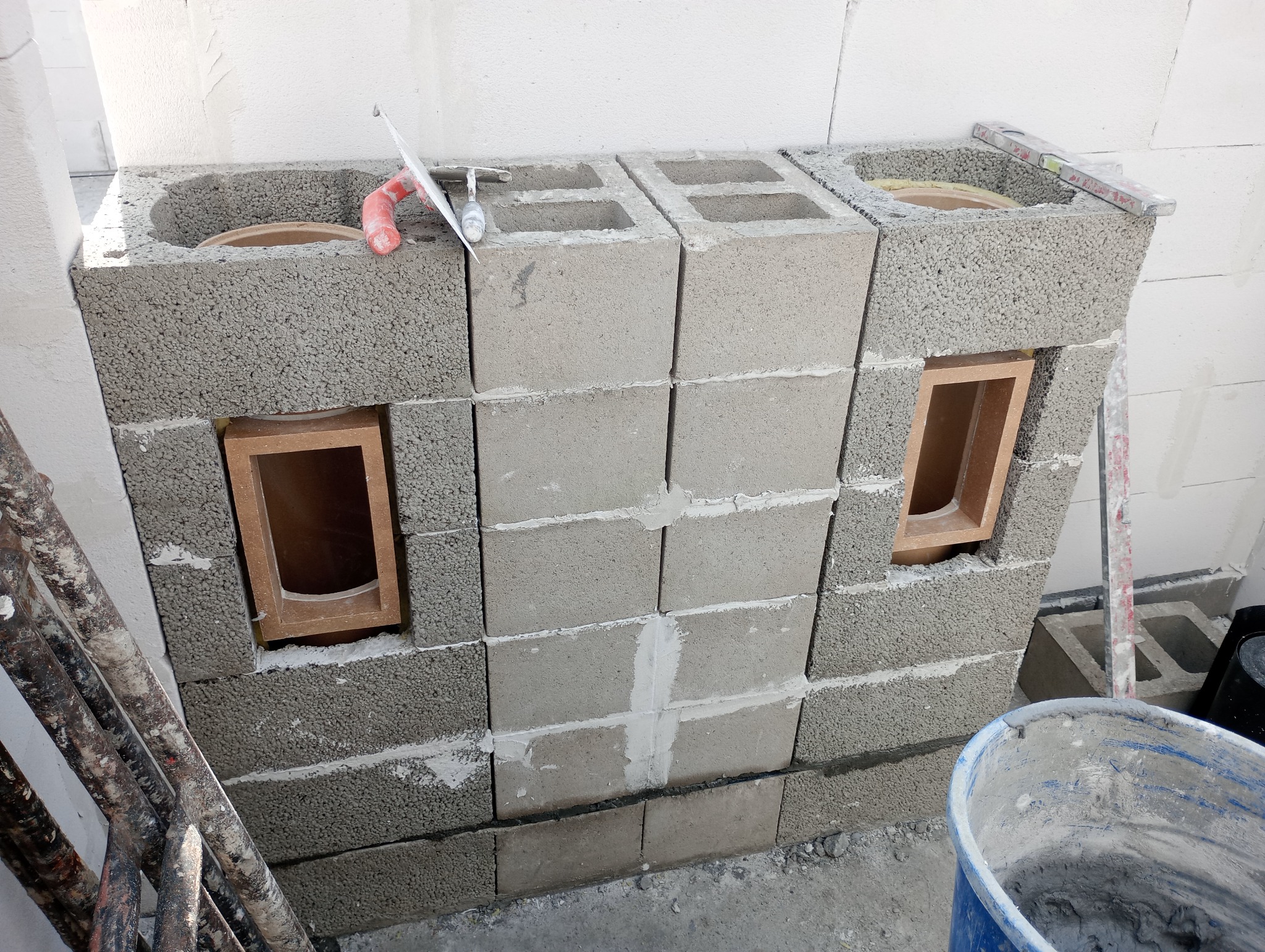 Wznoszenie konstrukcji z szarych bloczków betonowych z otworami, widoczne narzędzia murarskie i poziomica. Budowa kominka lub grilla w trakcie realizacji.