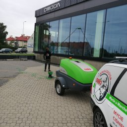 Mycie u Marchewy Michał Marchlewski - Pracownik myje przeszkloną elewację budynku z użyciem specjalistycznego sprzętu Dibo. W tle zaparkowany samochód z napisem 'Mycie Ciśnieniowe'.