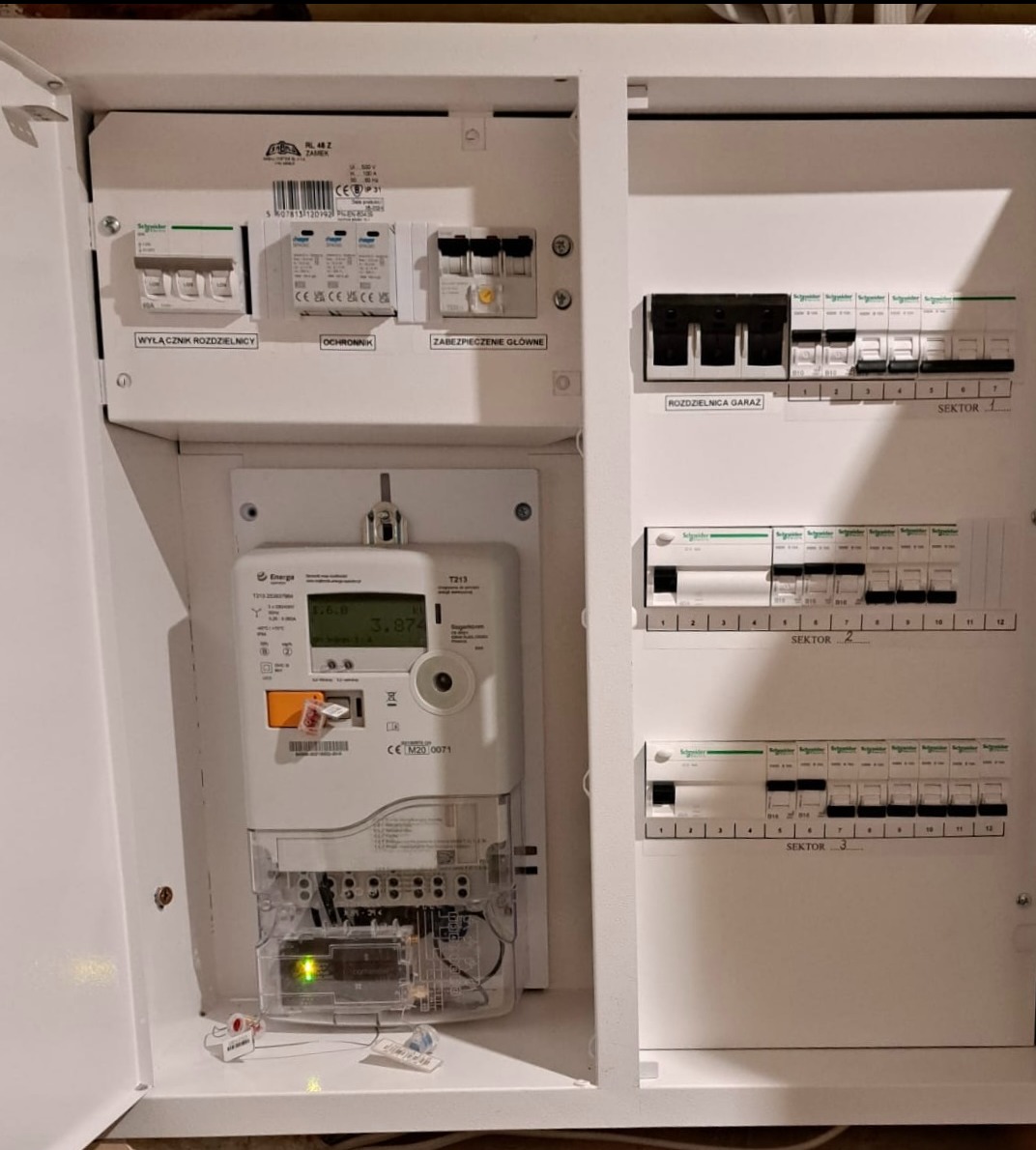 Biała skrzynka rozdzielcza z licznikiem energii elektrycznej, wyłącznikami i zabezpieczeniami. Widoczne oznaczenia sektorów i parametrów na liczniku.